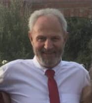 Man with short gray hair and beard wearing a white shirt and red tie, standing outdoors in a garden setting.