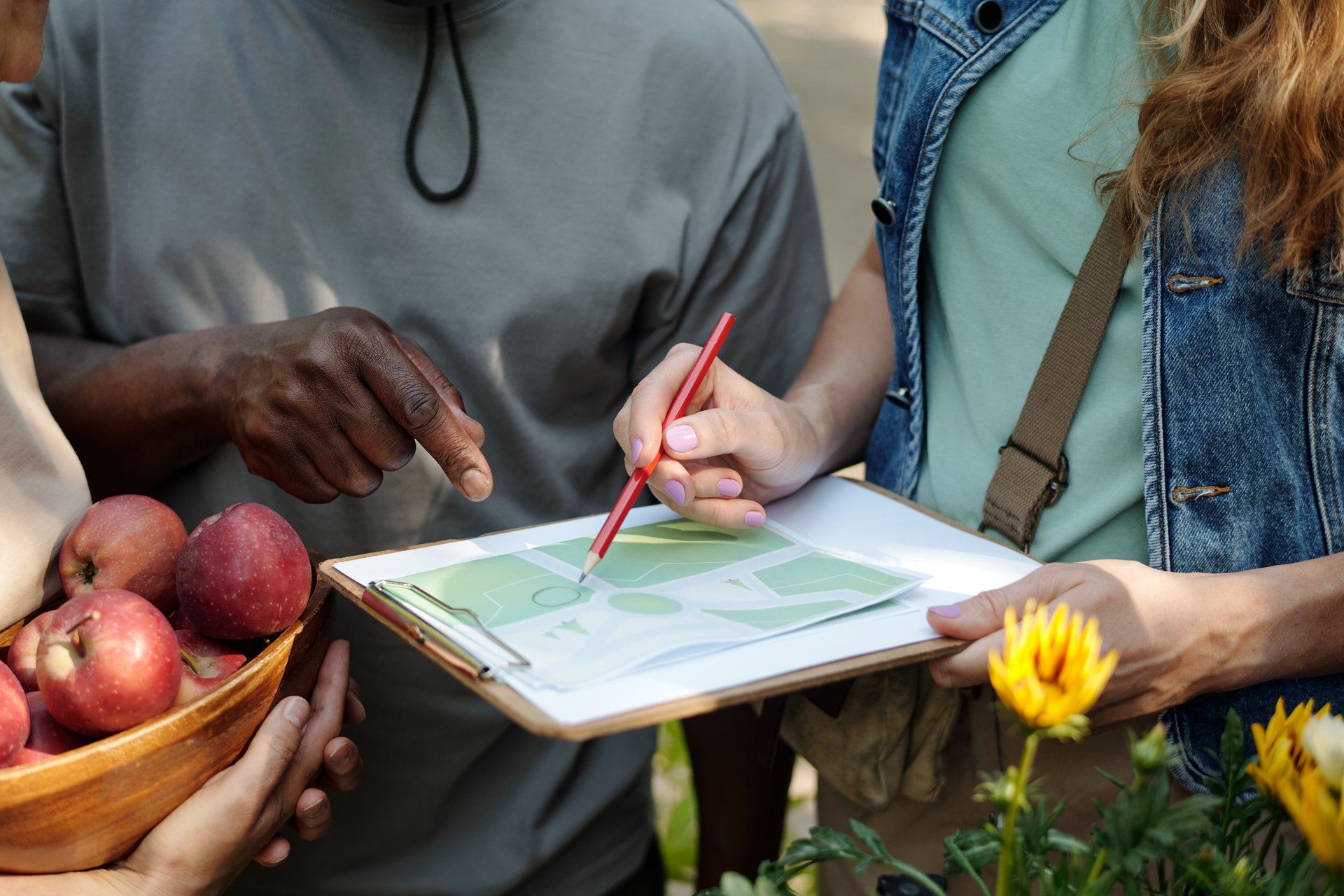 Young adult Caucasian woman holding clipboard with map, discussing directions with young adult Black man while another holding bowl of apples standing nearby, outdoor setting