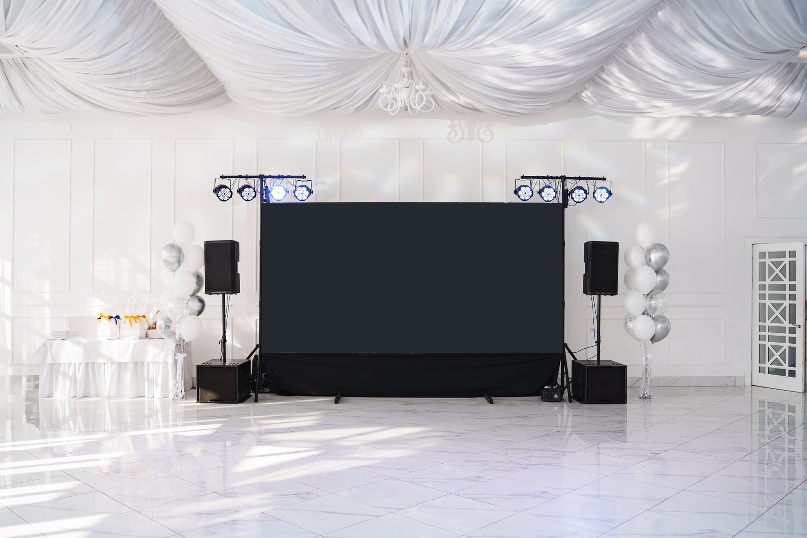The elegant venue features a large blank screen, professional lighting, and decorations. The space is designed for hosting events with a modern ambiance, ideal for presentations or celebrations.