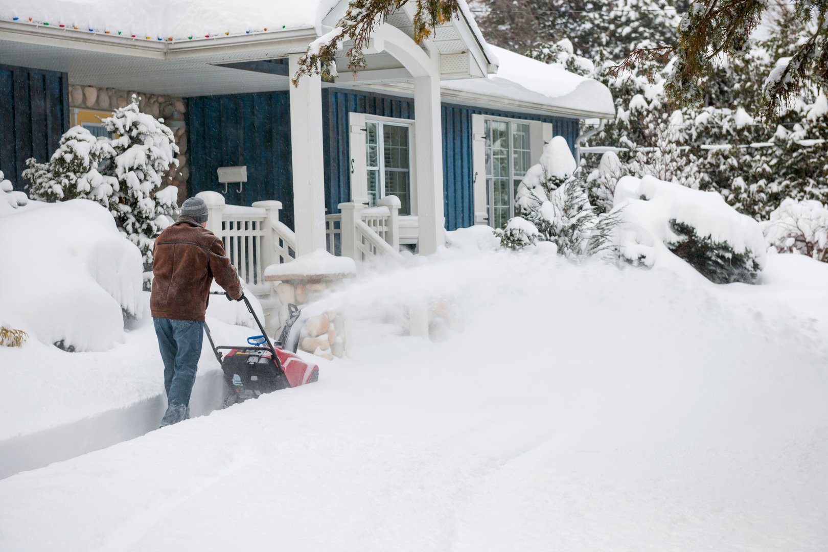 Person using a snow blower to clear a residential driveway covered in snow, with a blue house and snowy landscape in the background, illustrating snow removal services in Anchorage.