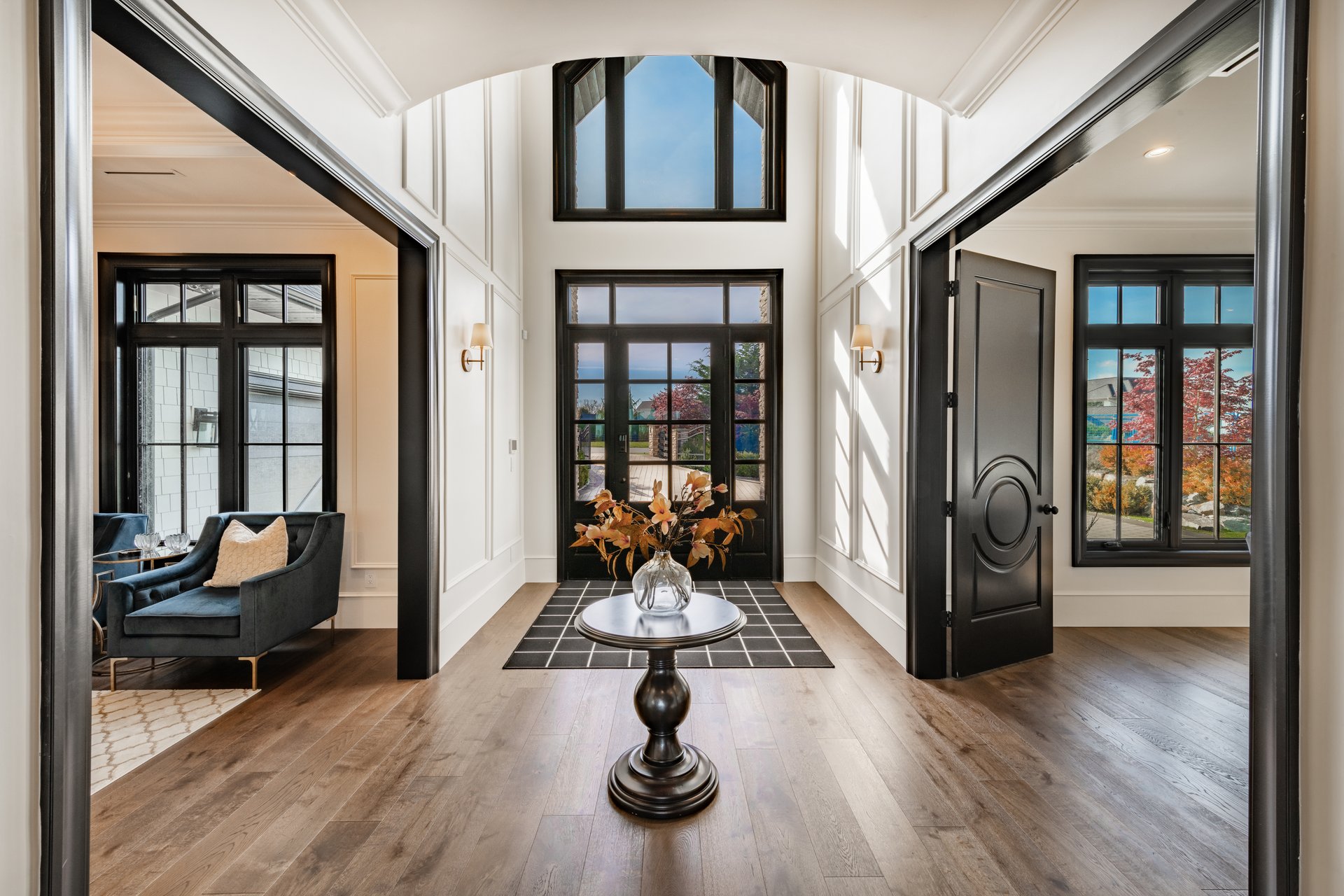 Beautiful large and luxurious estate home interior front door entry hallway foyer with large glass and black wood door expansive entry with a round table containing an arrangement of dried flowers in a bouquet. The view from the front is to a home office and a casual sitting room with blue velvet sofas. large wooden barn doors lead to another room there is a staircase and steps going up.