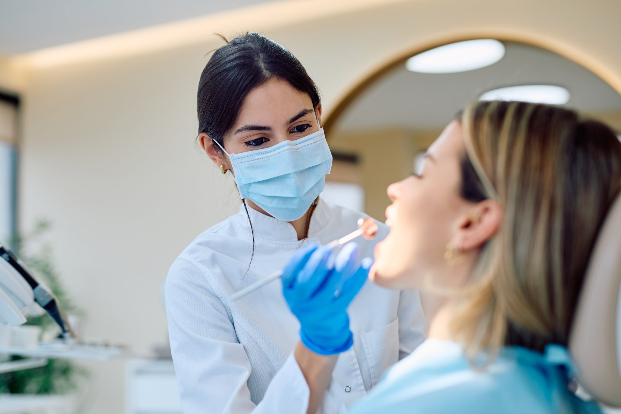 Female dentist wearing mask and gloves performing an oral examination on a woman in a modern dental clinic. Dental care concept