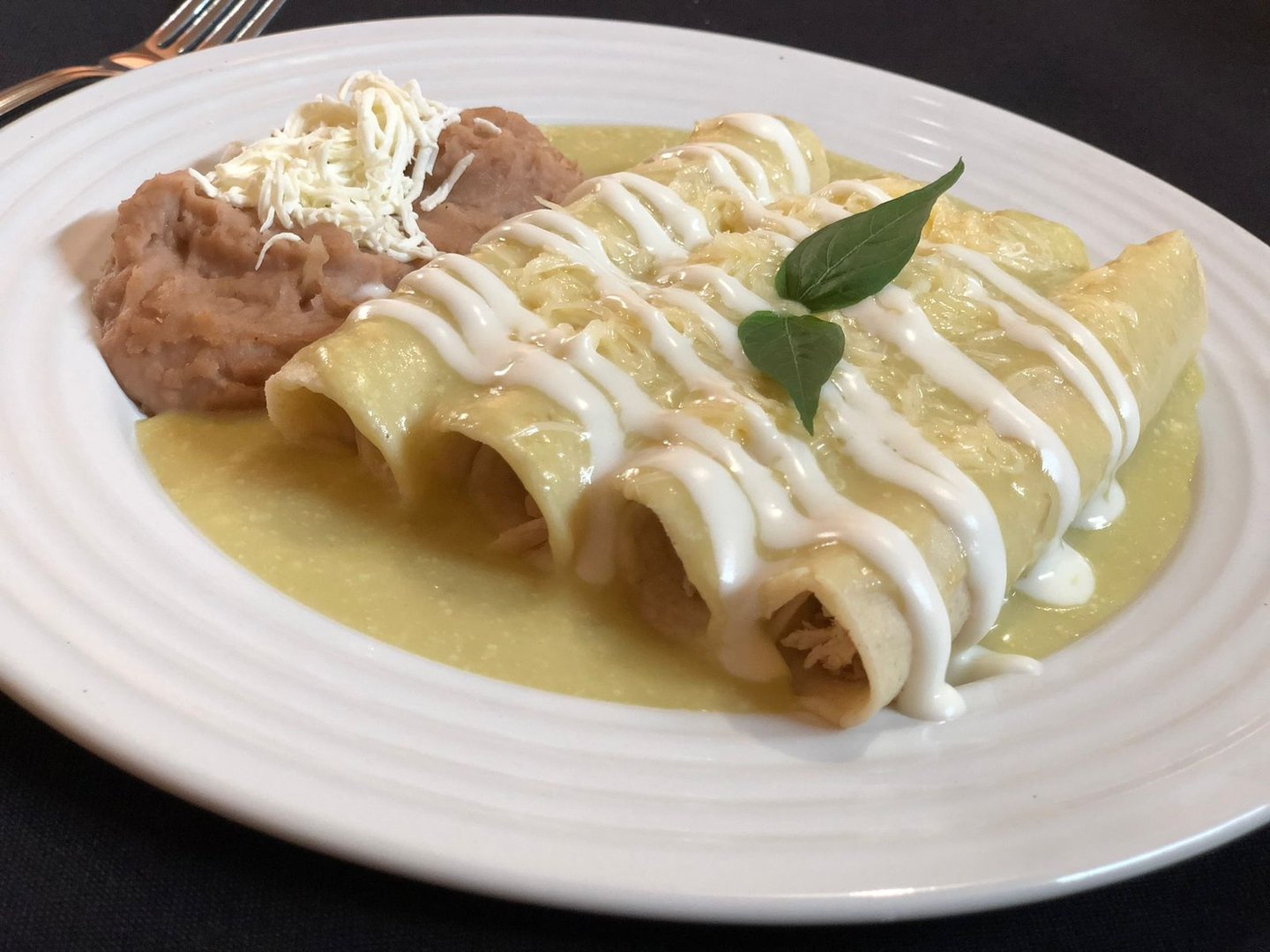 Plate of enchiladas with green sauce, cream drizzle, and refried beans topped with cheese, garnished with a herb leaf.
