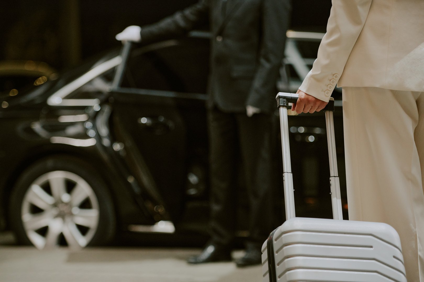 Caucasian middle aged man holding suitcase standing near luxury car while professional driver in uniform opening door, business travel concept, partial body view, no faces visible