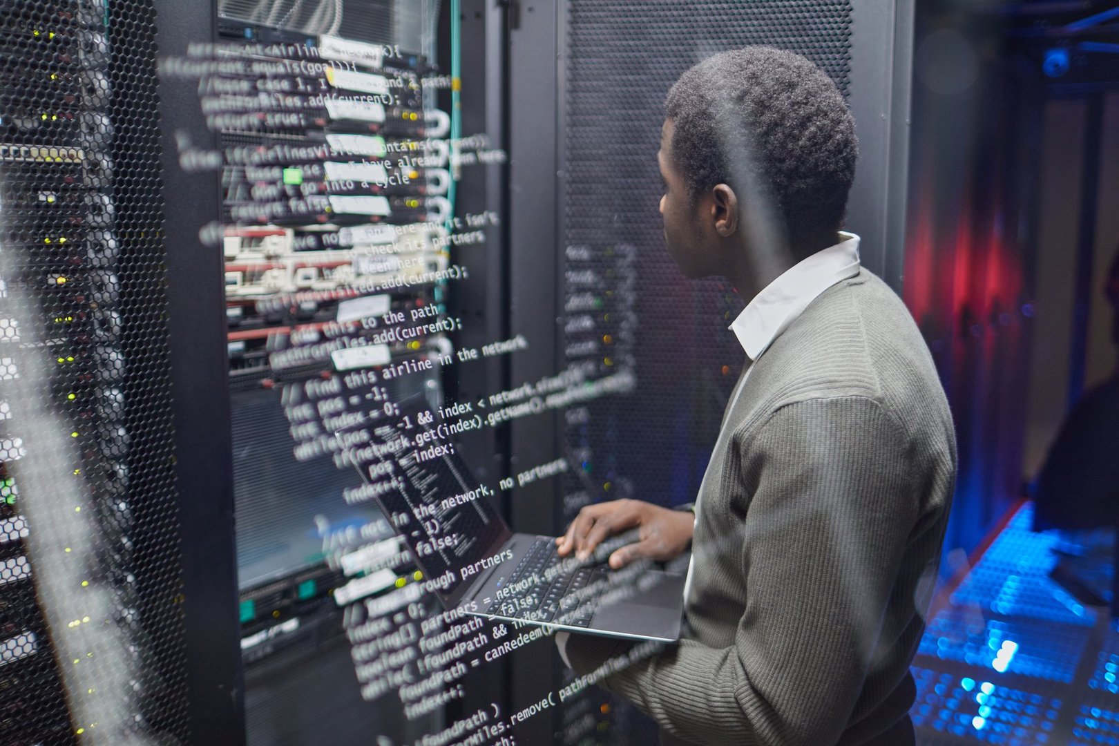 African American technician checking data systems in modern data center, monitoring status on laptop among servers and cables, ensuring optimal performance and security