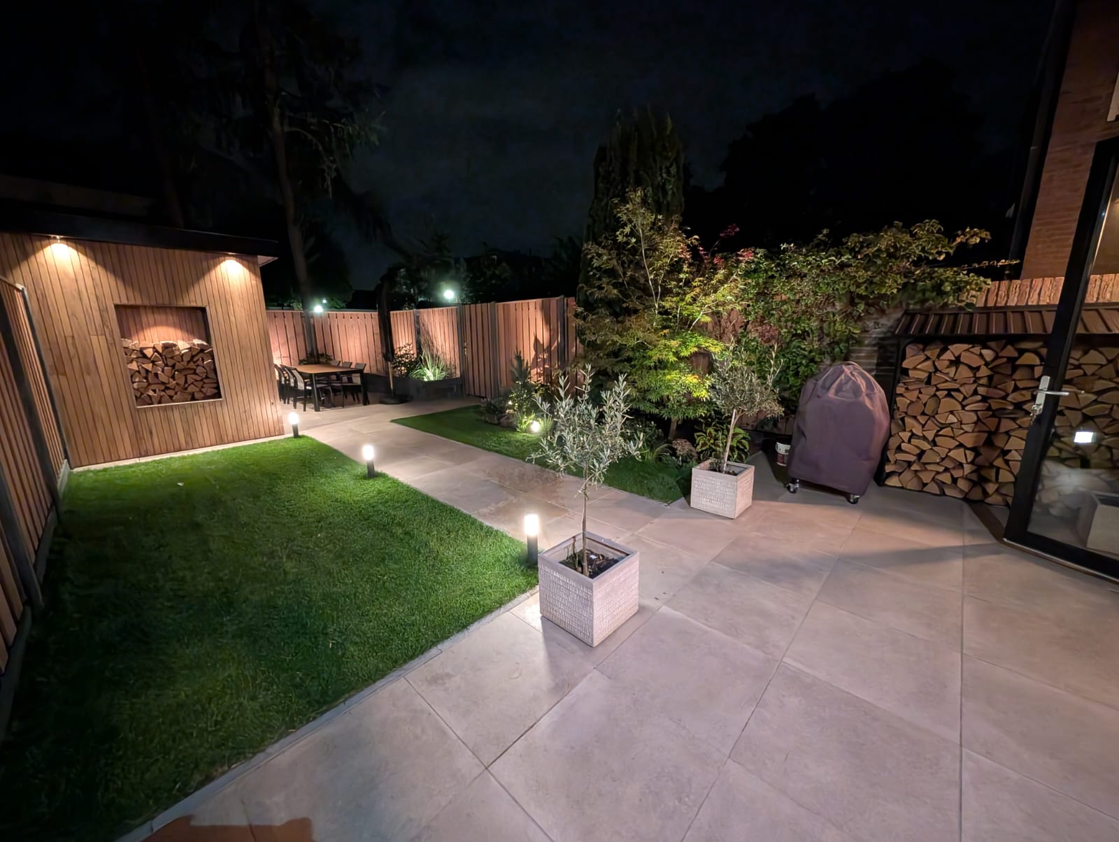 Nighttime garden with patio, potted plants, stacks of firewood, and soft lighting illuminating the pathway.