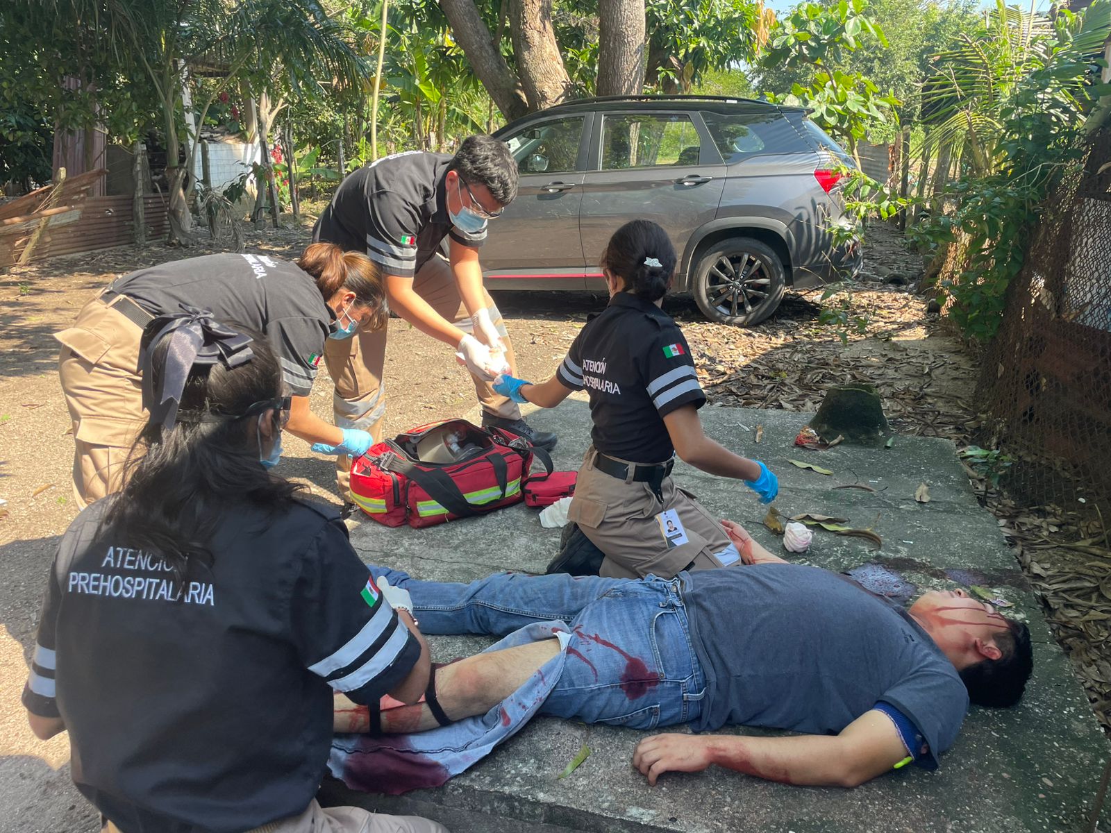 Emergency responders treat an injured person lying on the ground next to a parked car. Medical supplies are visible.