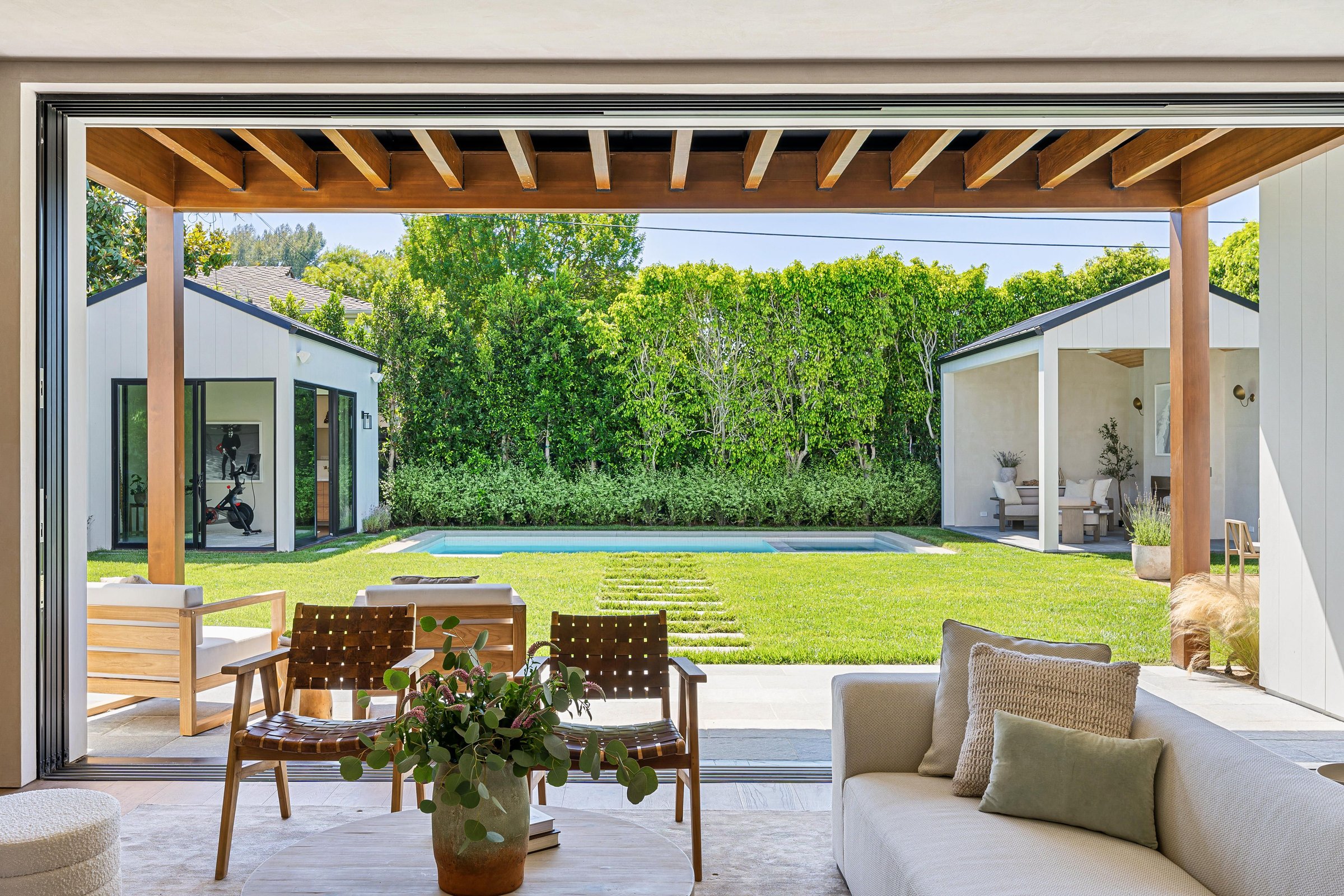 Modern open living space with view of a lush backyard and pool, featuring wooden beams and contemporary furniture.