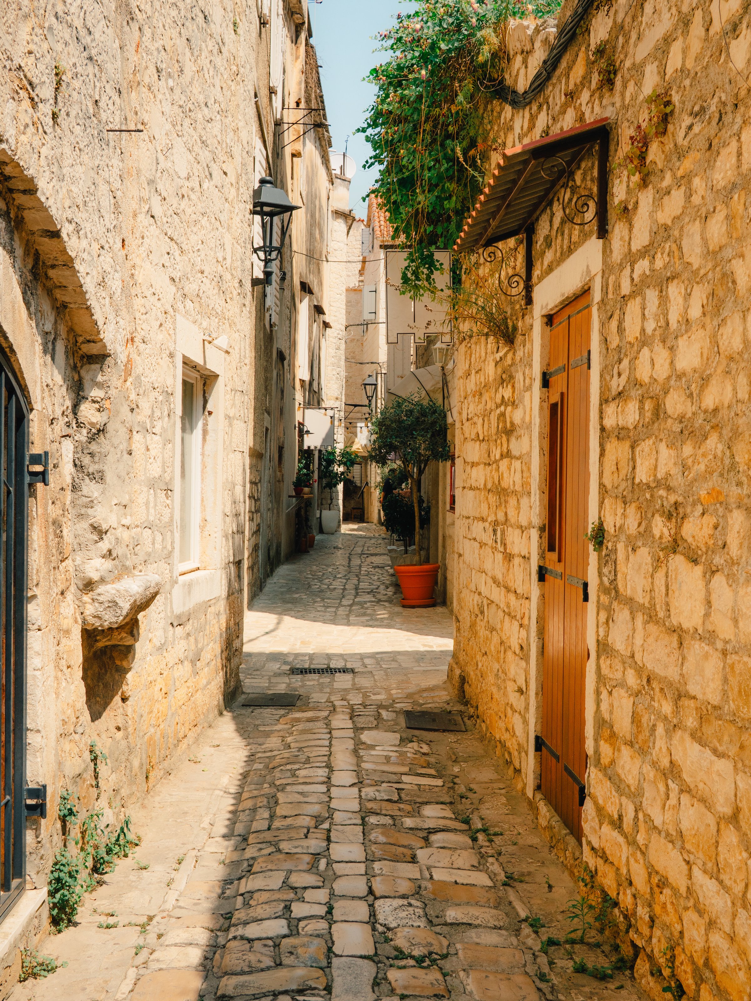 Historic cobblestone street in Ulica Matije Gupca, Trogir, Croatia, lined with stone buildings, arched doorways, lanterns, and lush greenery in the heart of the old town.