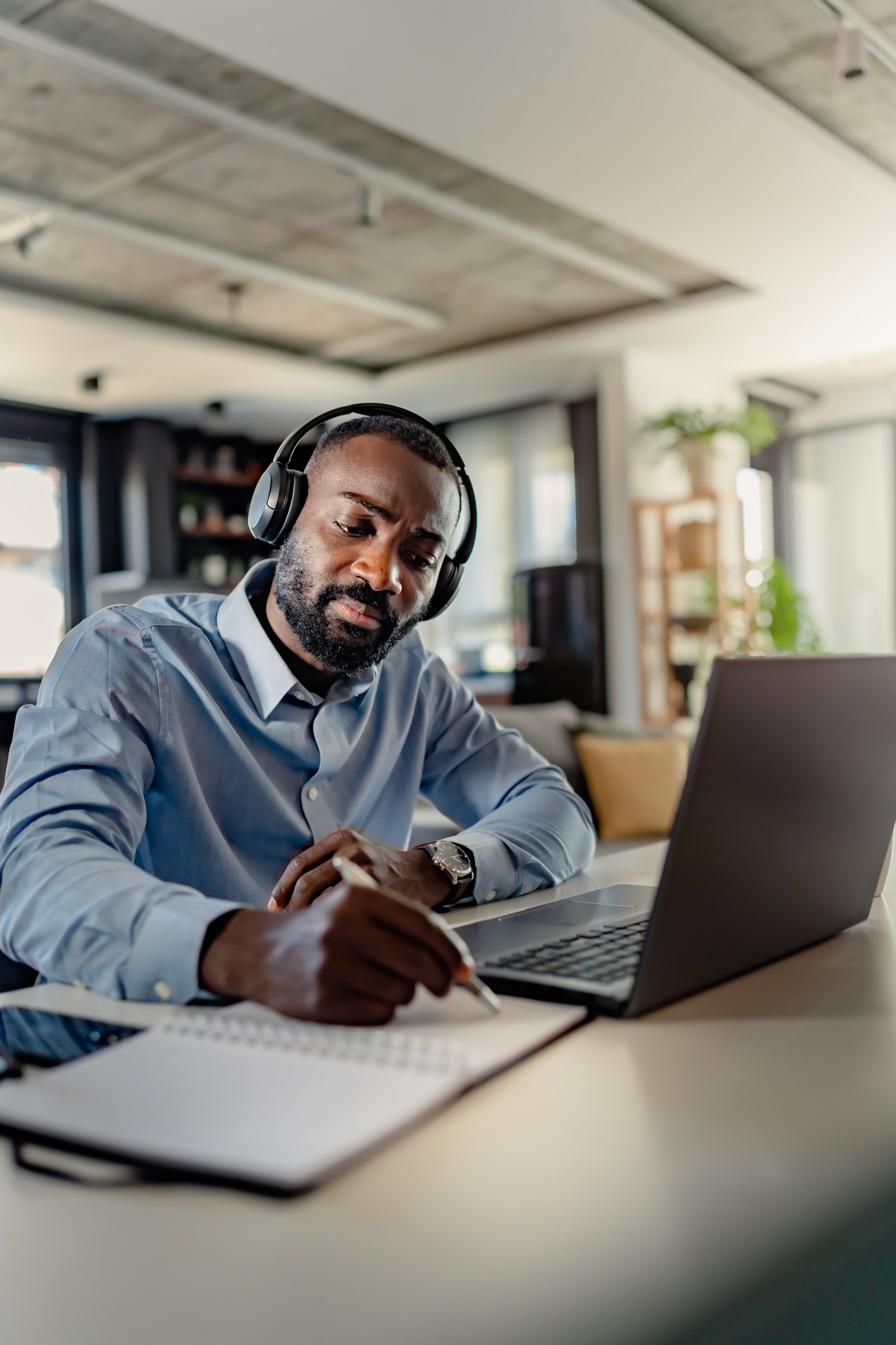 African-American man wearing headphones, taking notes while working on a laptop from home, focused and engaged in a modern workspace.