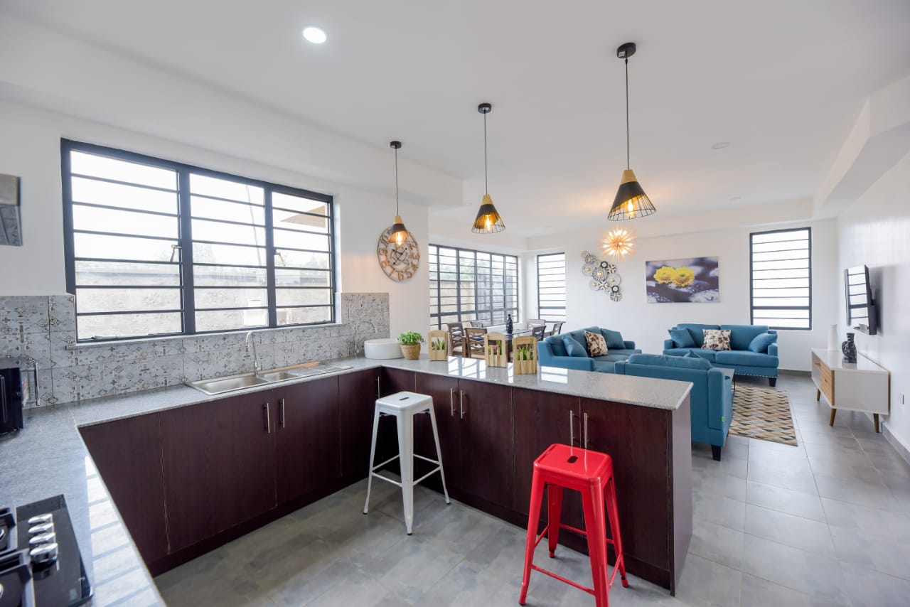 Modern open-plan living area with kitchen, dining, and lounge; features blue sofas, pendant lights, and large windows.