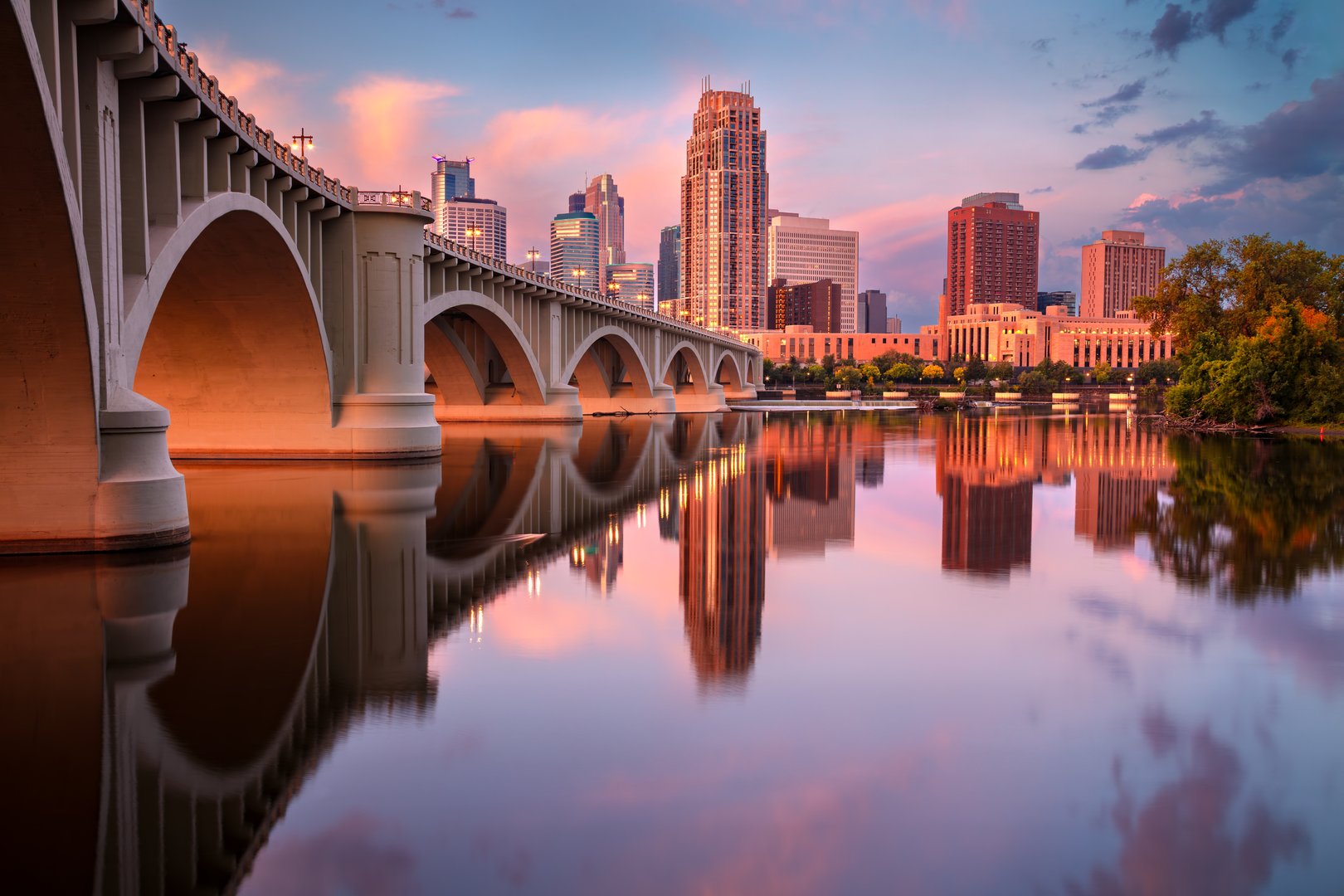 Minneapolis, Minnesota, at sunrise features the city skyline reflected in a calm Mississippi River. The cityscape showcases modern high-rise buildings under a pink and purple sky. In the foreground, an arched bridge spans the river, its structure highlighted by soft morning light. Trees with autumn foliage line the riverbanks, enhancing the scene with warm colors. The water's surface creates a mirror-like reflection of both the architecture and the sky, adding to the picturesque setting.