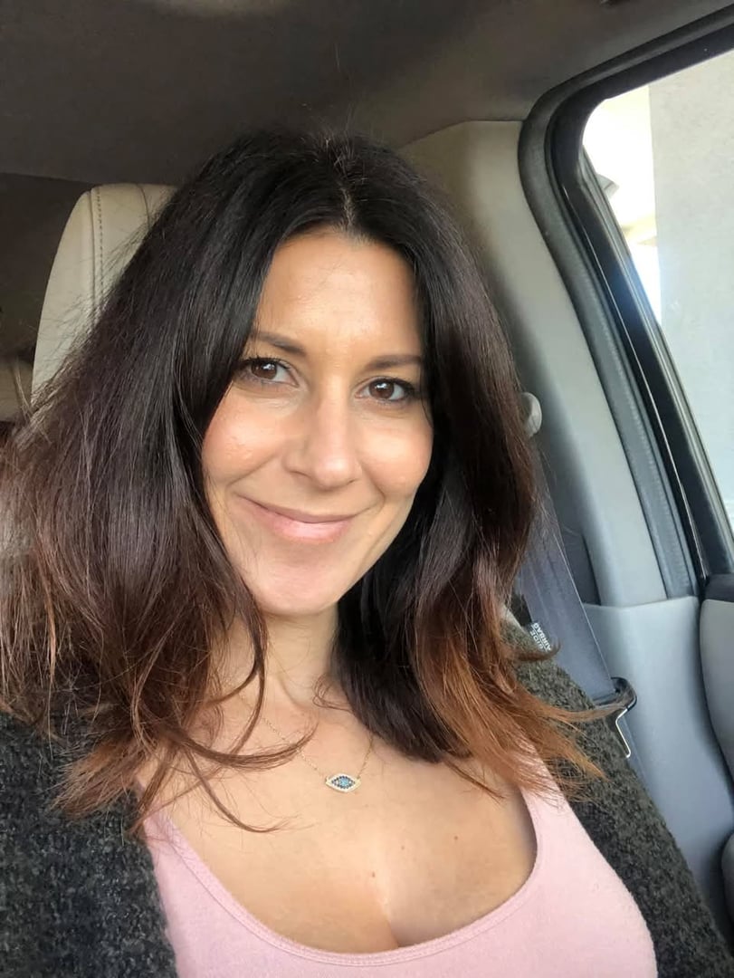 Woman with dark hair smiling in a car, wearing a pink top and a necklace with an eye pendant.