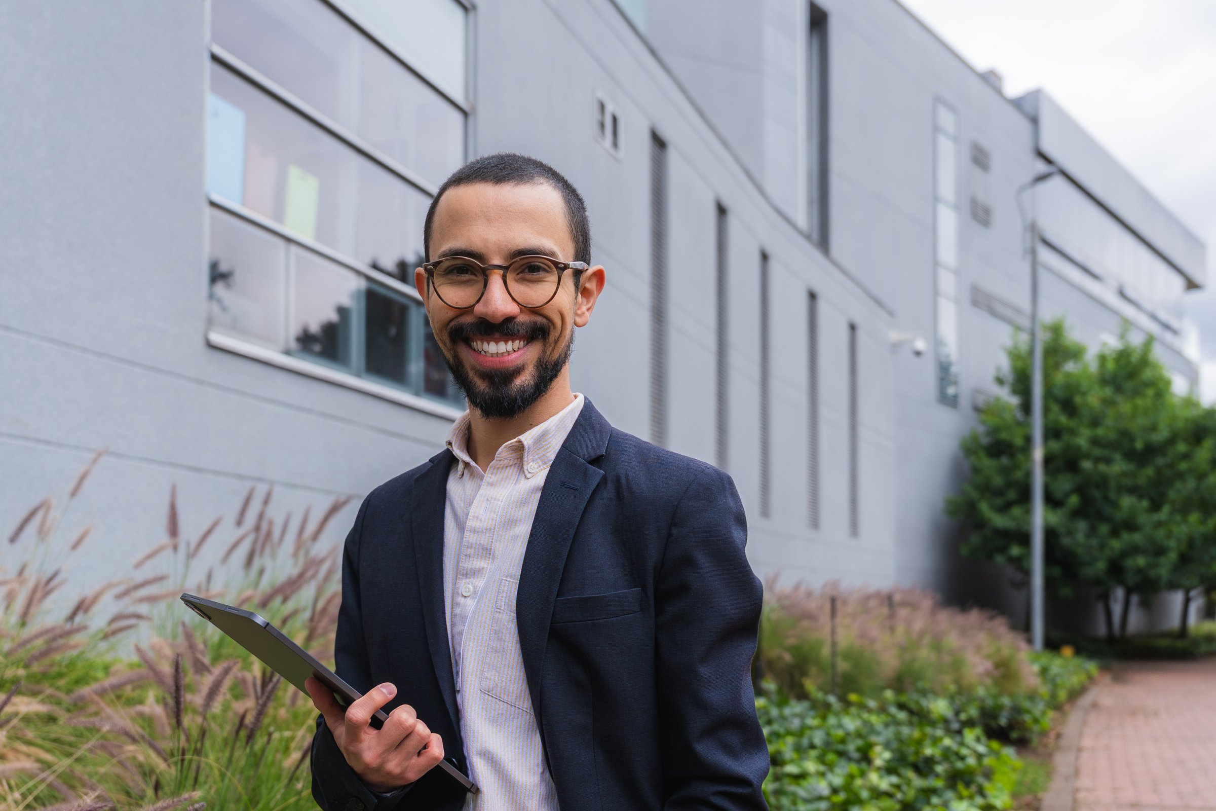 Young man standing outdoors, wearing glasses and a suit, holding a digital tablet