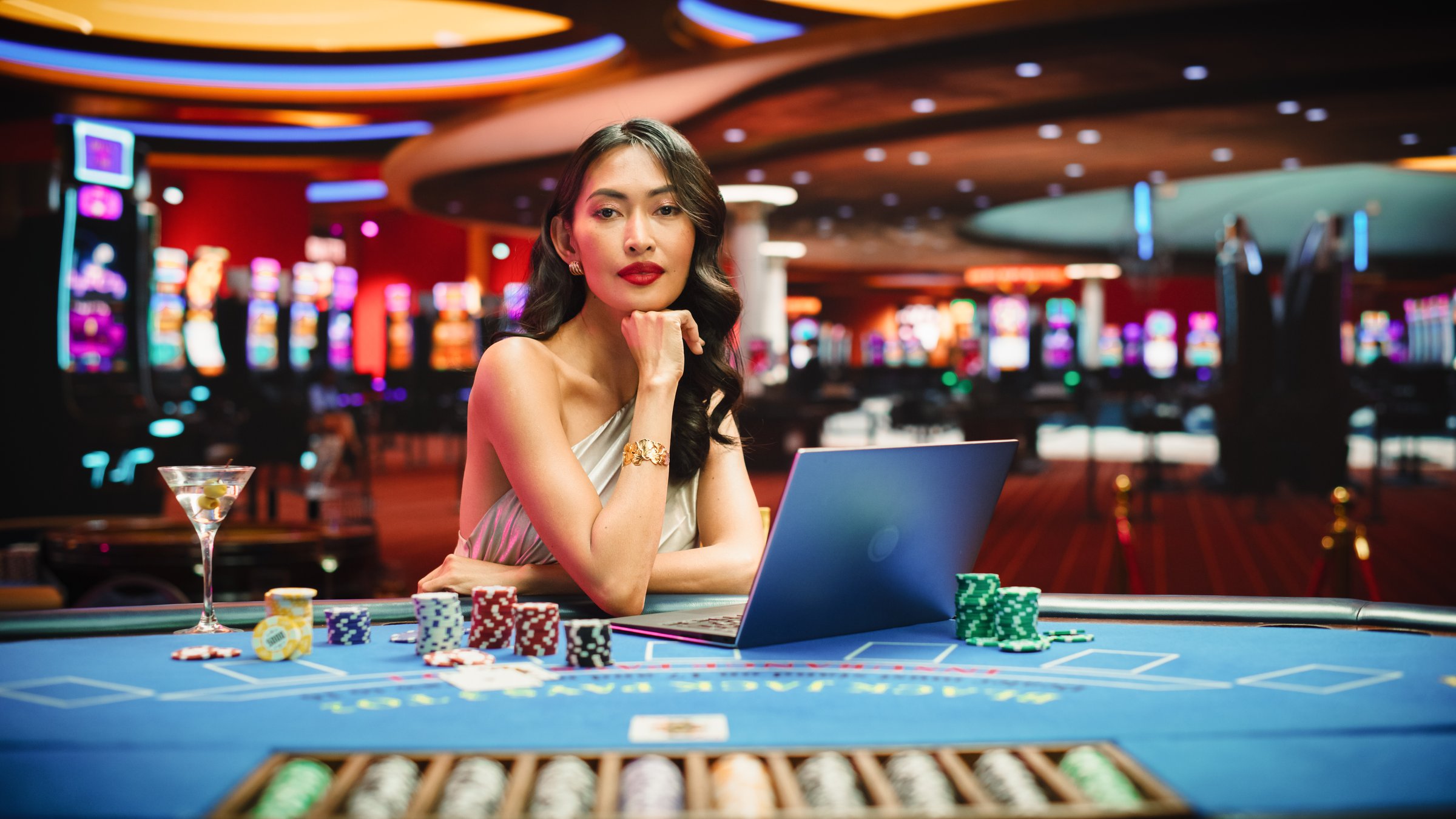 Glamorous Asian Woman at Casino Table, Engaged in Online Betting Games on Laptop, After Winning Bet she Turns and Looks Elegantly at the Camera. Glamorous Successful Female Playing Online Blackjack