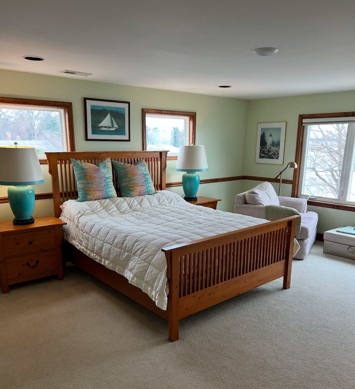 Bedroom with a wooden bed, colorful pillows, turquoise lamps, art on the walls, and a white armchair by the window.