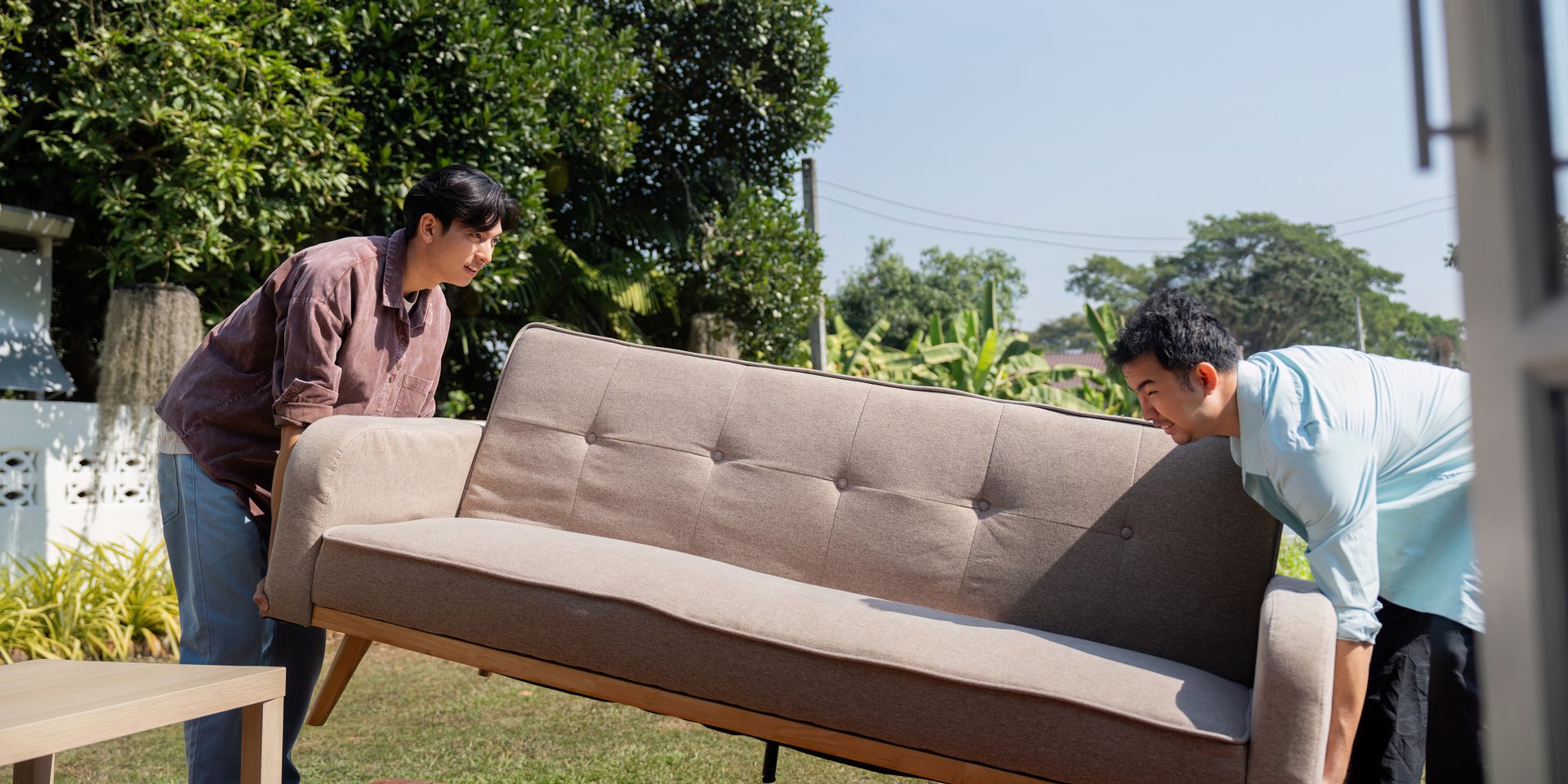 Two men, a couple, working together to move a sofa into their new home, surrounded by greenery in a beautiful outdoor setting.
