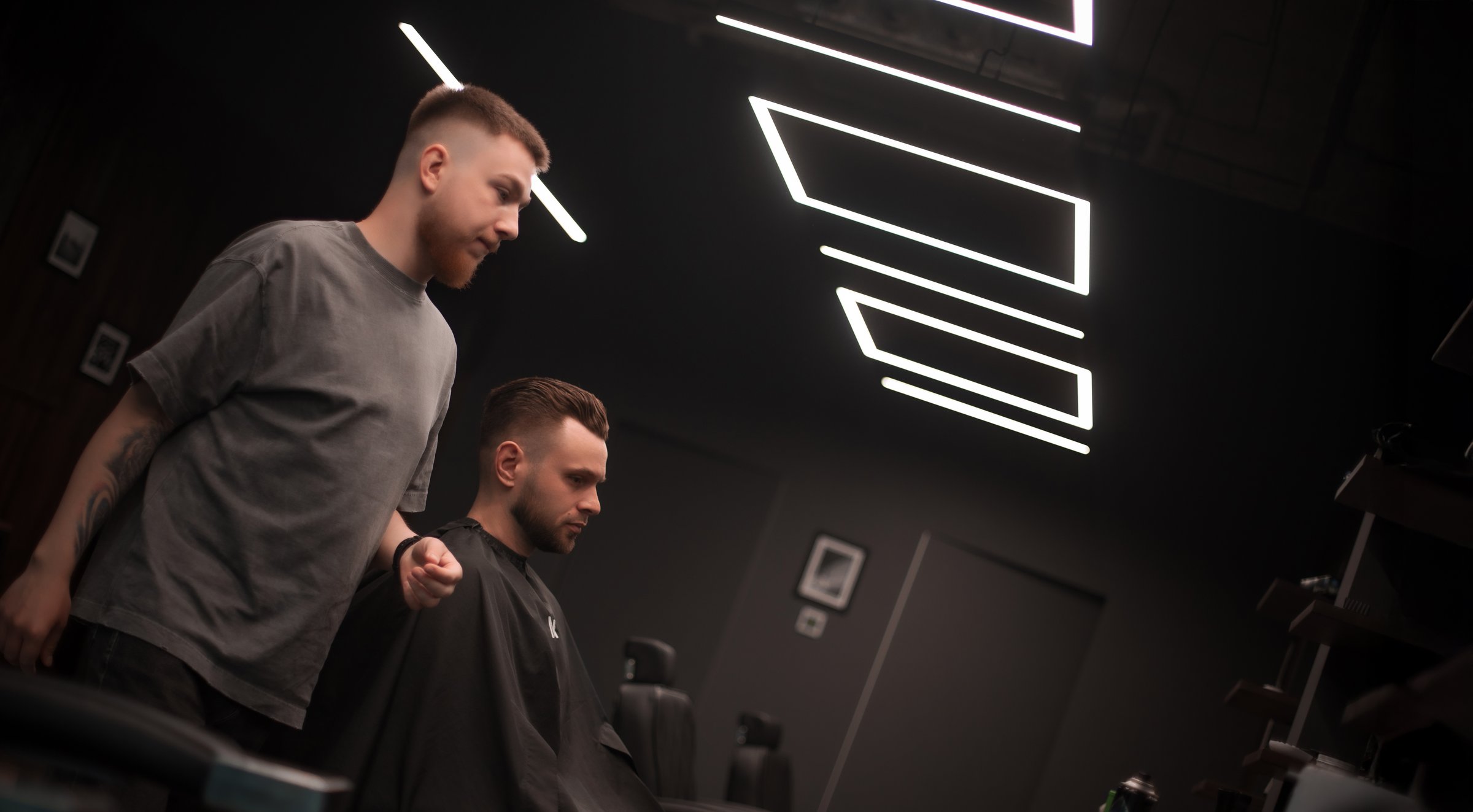 Barber and client in modern barbershop. The barber stands next to a seated man wearing a cape, preparing for a haircut under stylish geometric ceiling lights. Stylish grooming scene.