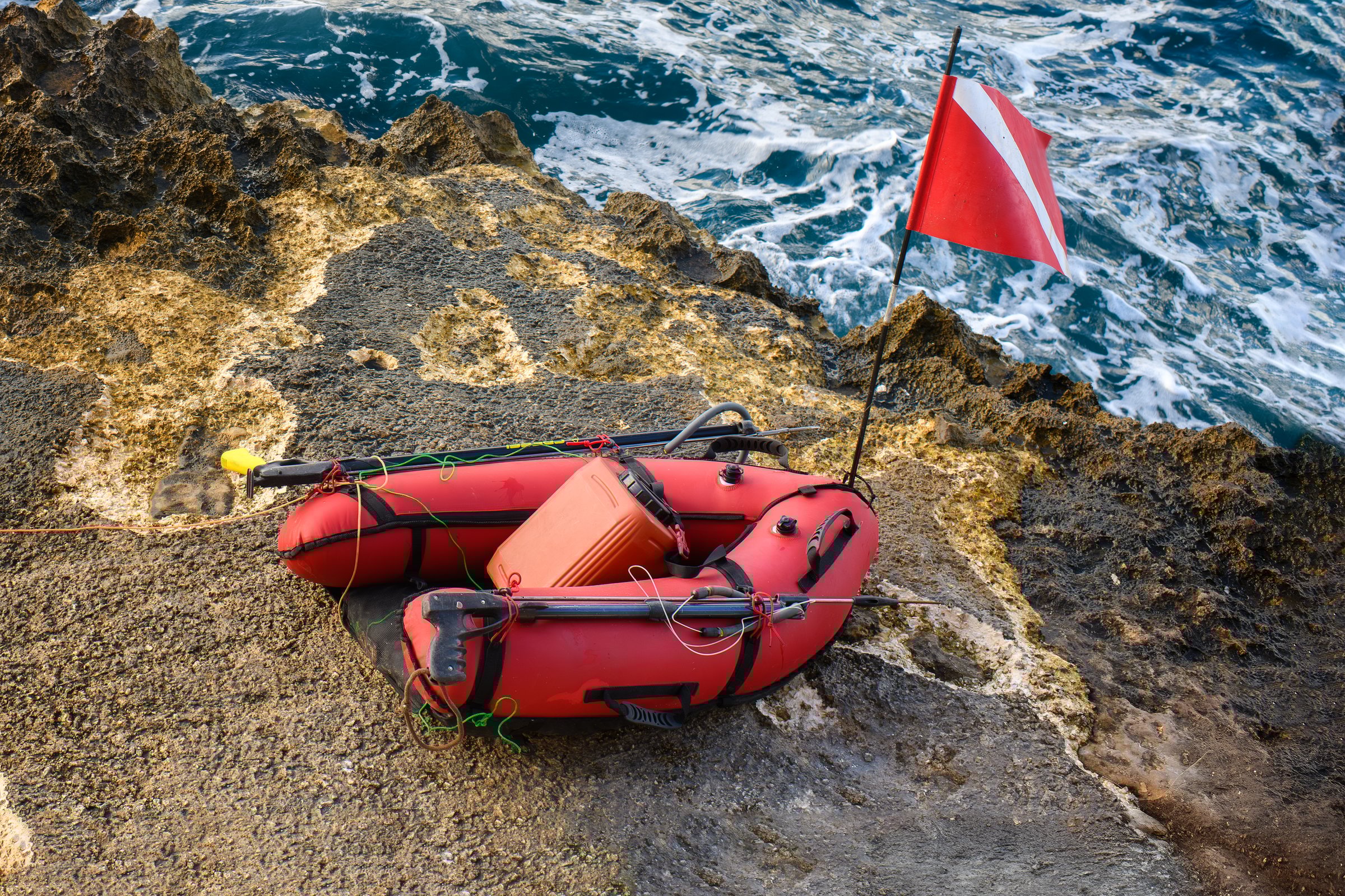 Red diver down float with a safety flag, spearguns, and spearfishing gear rests on the rocky shore beside the choppy sea.