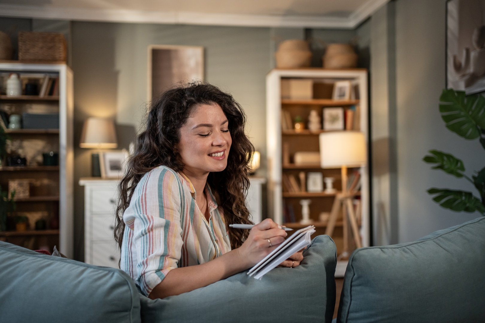 Young woman with long brown hair sits comfortably on her sofa at home, smiling as she writes in a notebook, enjoying a quiet moment of reflection and productivity