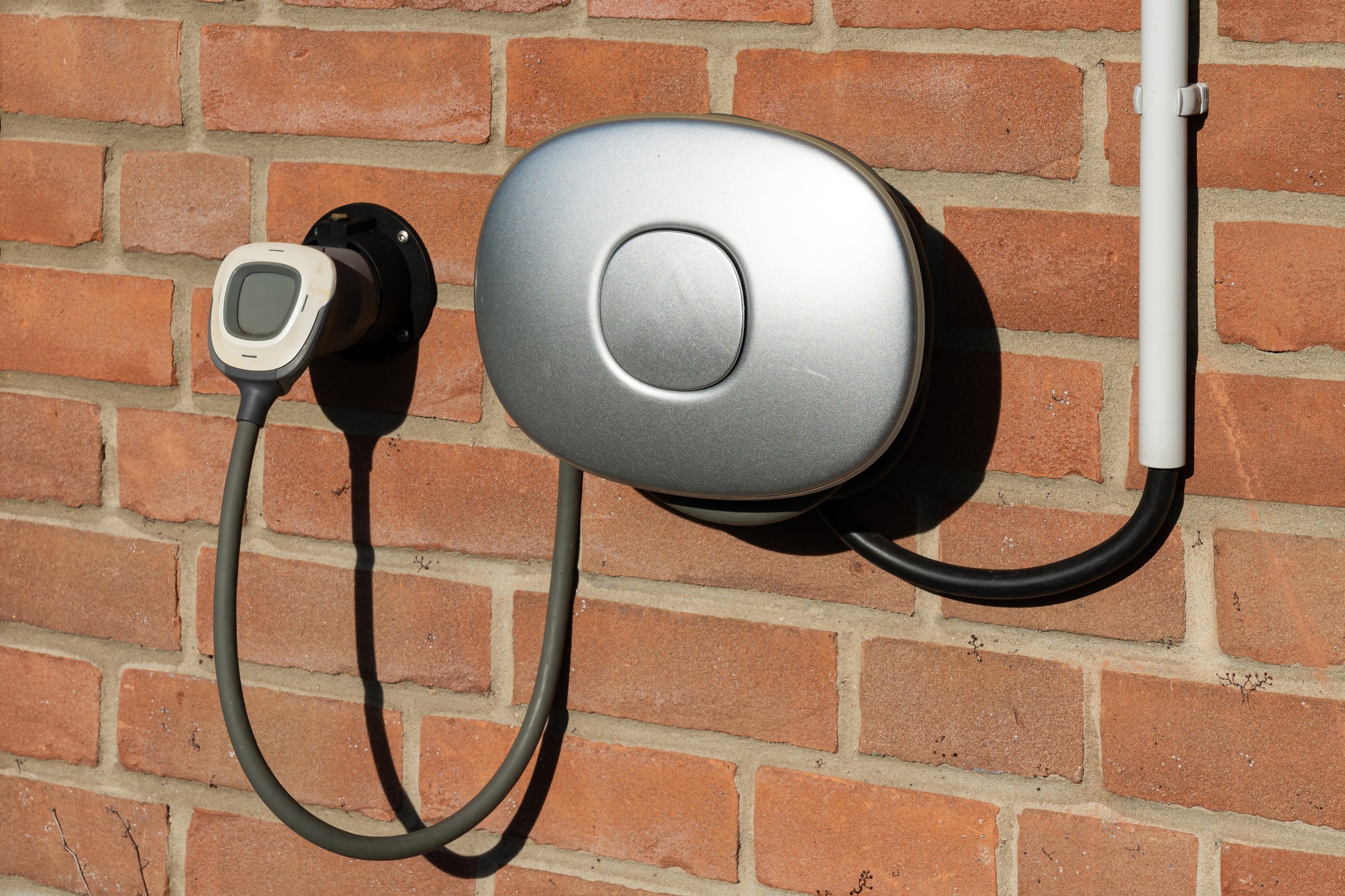 A modern electric vehicle charging station is mounted on a brick wall, ready for use. The design features a sleek, rounded charger attached to a durable cable.