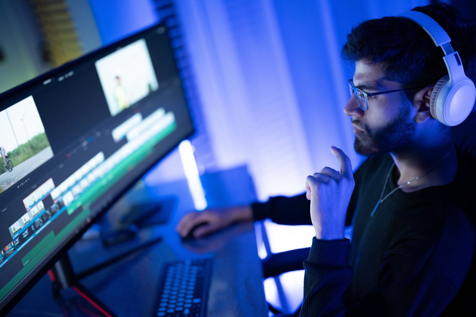 A young man focuses intently on his dual monitor setup, editing video content in a dimly lit room decorated with ambient blue lights. His thoughtful expression reveals concentration.
