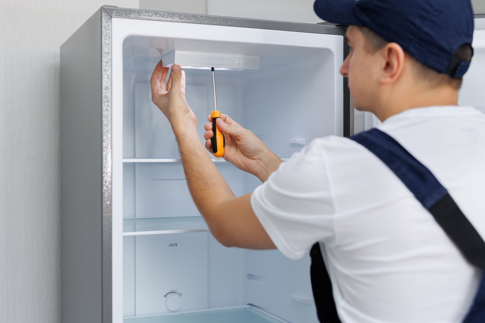 Man in a uniform repairs the light in the refrigerator with a screwdriver. Replacing the light bulb