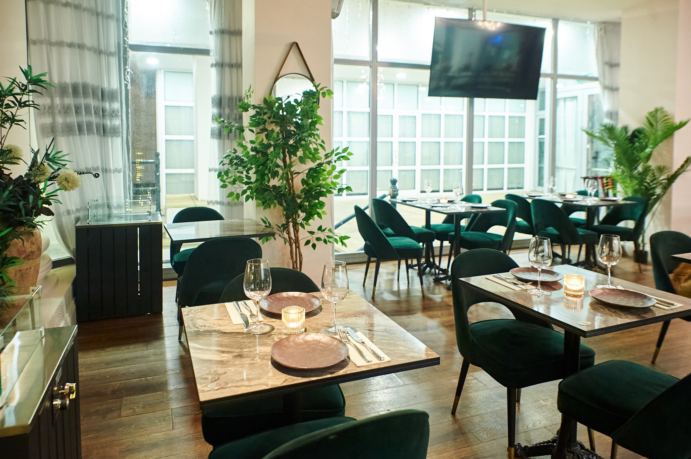 Elegant restaurant interior with green chairs, wooden tables set for dining, plants, and a wall-mounted TV.