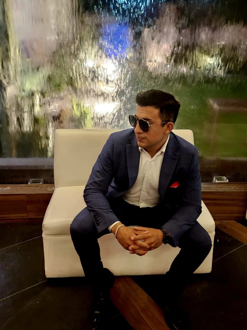 Man in a blue suit and sunglasses sits on a white couch in front of a water feature, looking to the left.