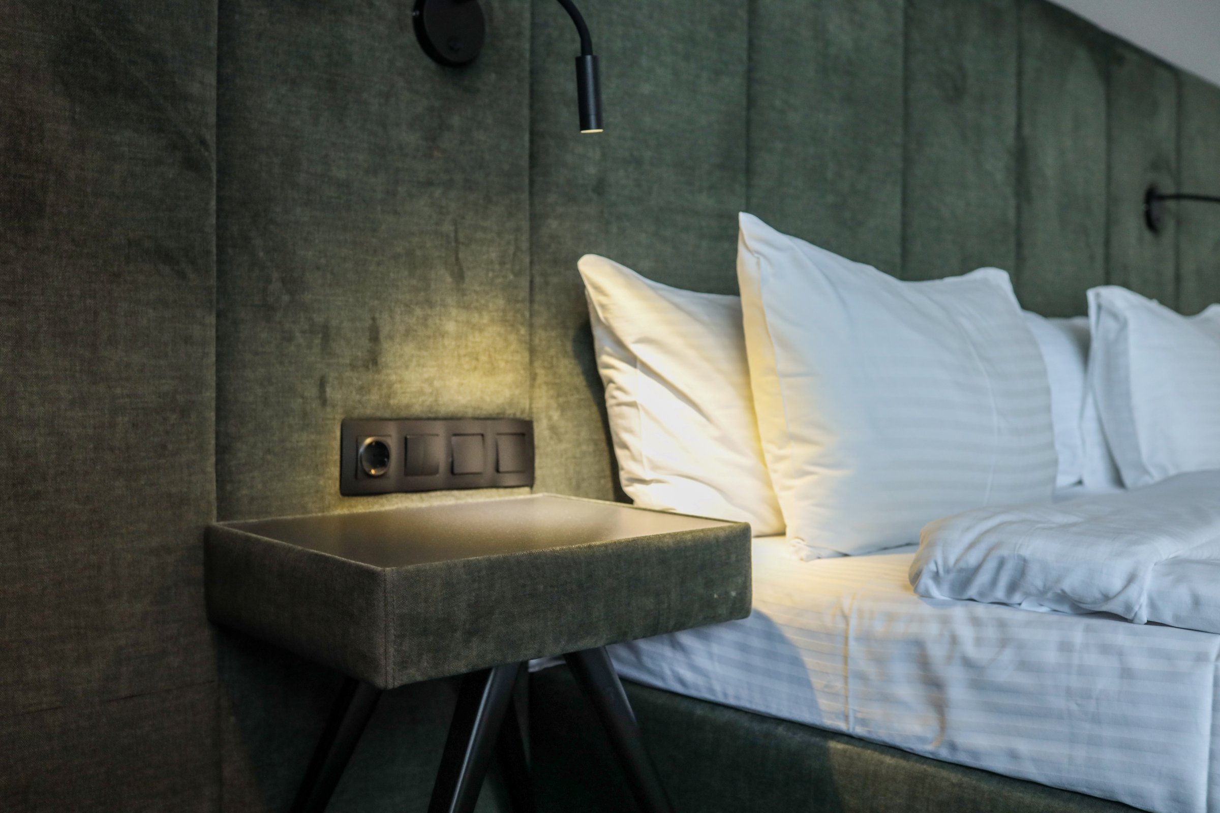 Close-up of modern hotel room interior with white pillows, clean bed, soft headboard and nightstand with built-in lighting. Cozy and minimalist design for comfortable stay.
