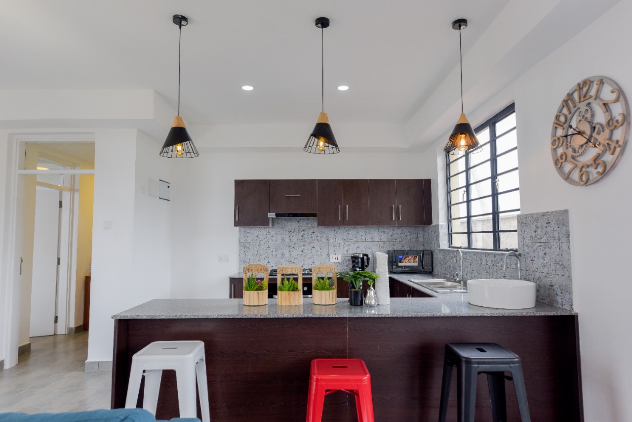 Modern kitchen with dark cabinets, bar counter with stools, hanging lights, and a wall clock.
