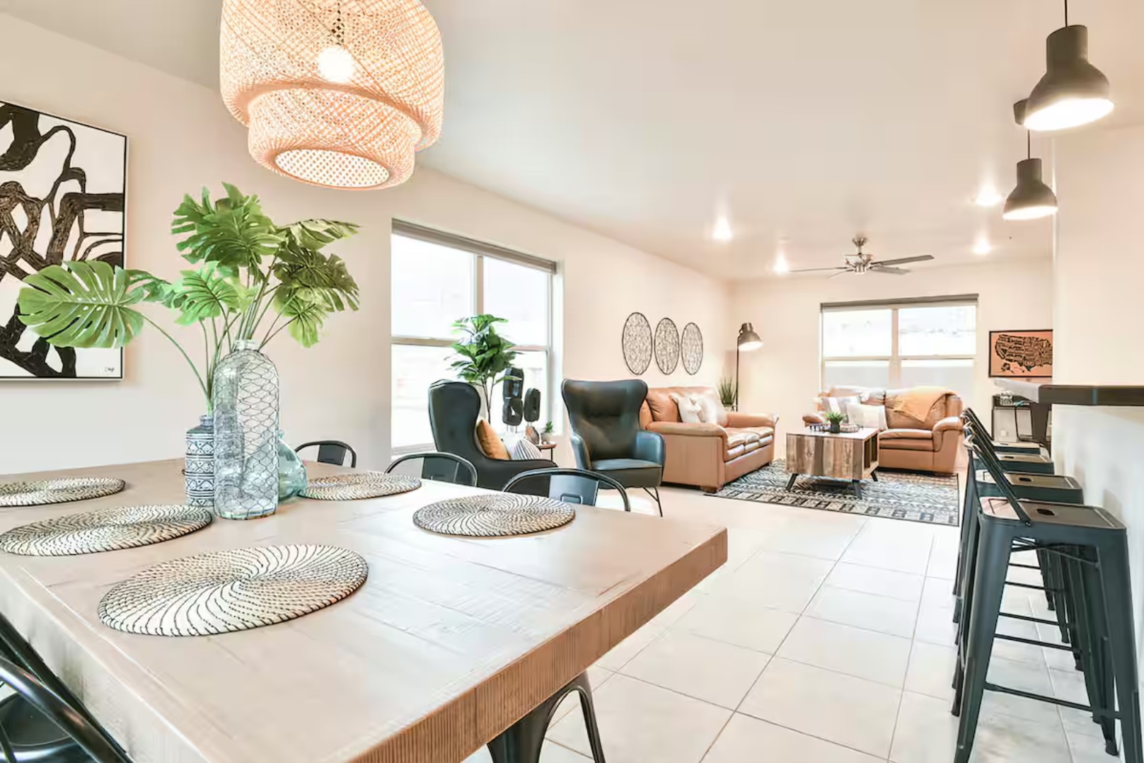 Modern open-plan living area with a dining table, potted plants, woven light fixture, and a cozy seating area in neutral tones.