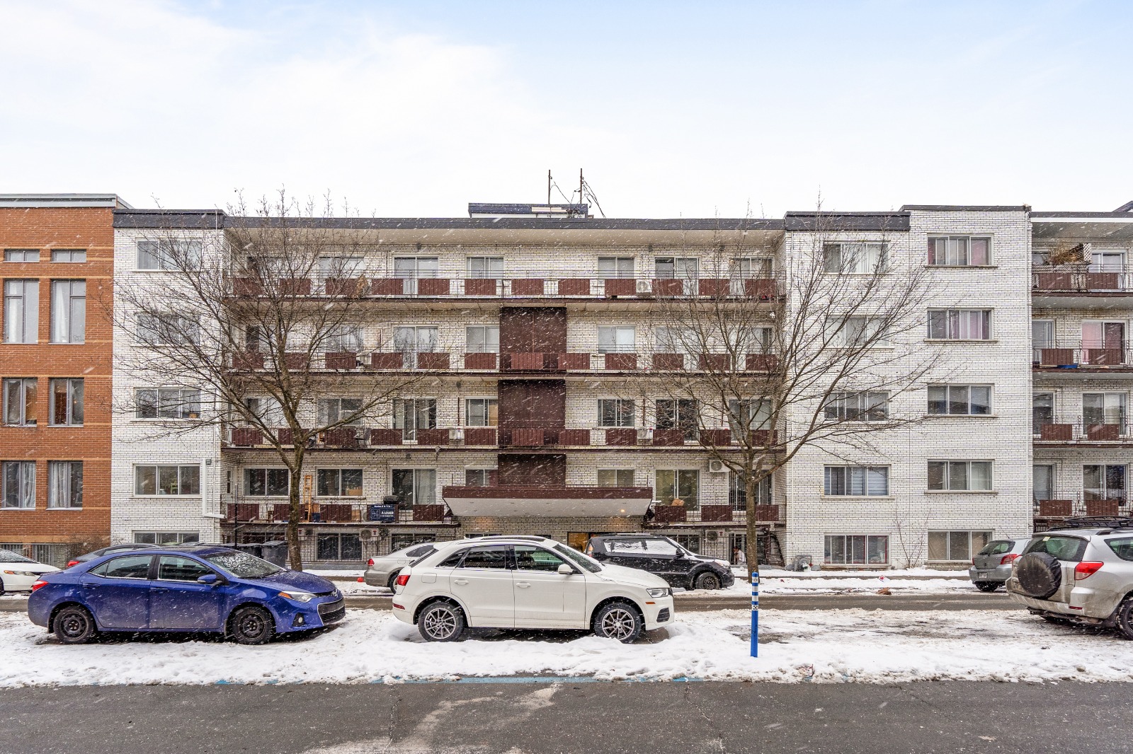 An apartment building in winter with snow on the ground and parked cars in front, including a blue and a white car.