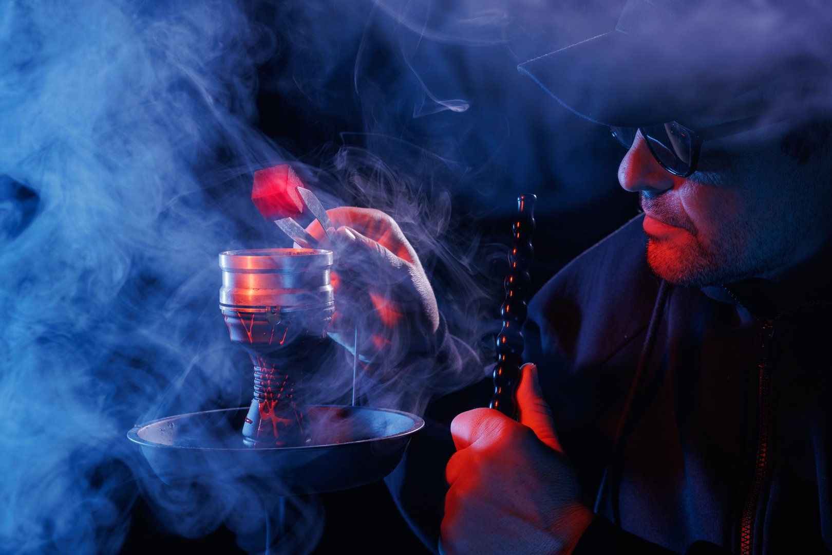 Man smoking hookah, adjusting hot charcoal with tongs in a smoky, moody atmosphere, illuminated by red and blue lighting