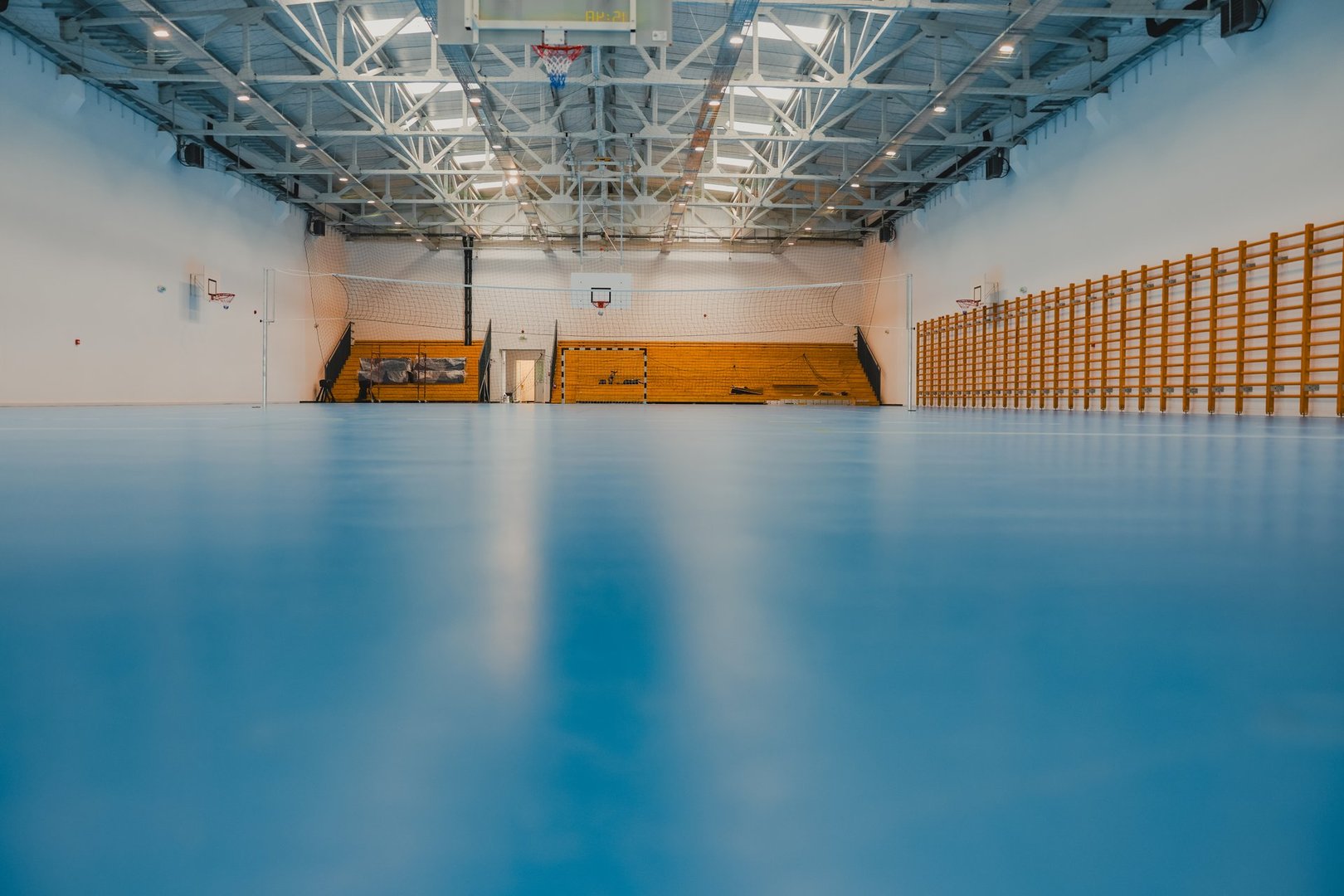 Wide shot of an empty sports hall with blue epoxy flooring, bleachers, basketball hoops, and wall bars