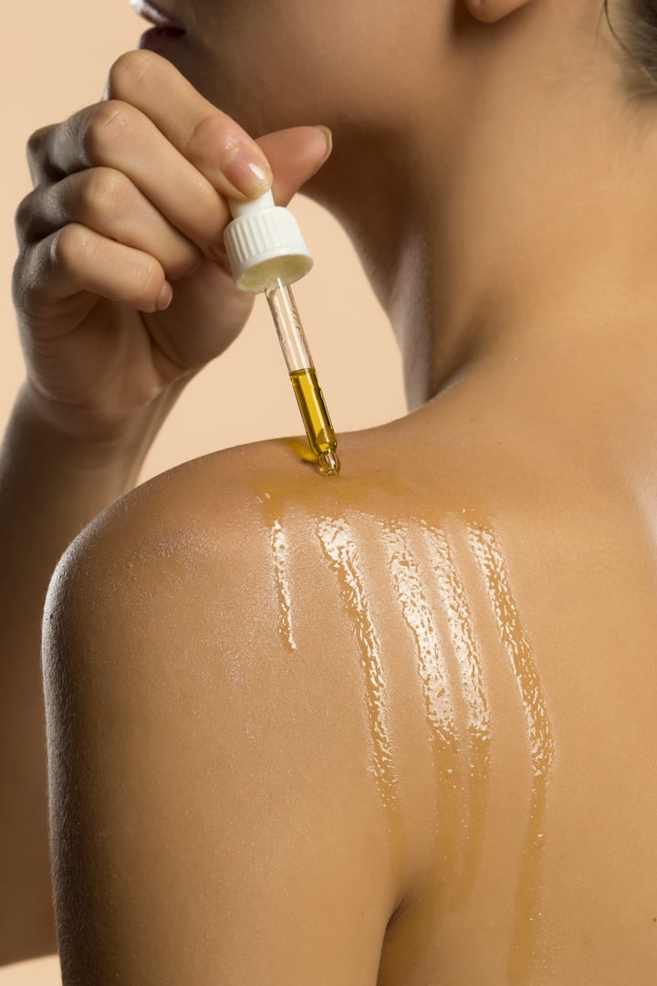 Close-up of woman applying olive oil to shoulder with dropper. Streaks of oil visible on skin, demonstrating natural skincare treatment. Neutral studio background.