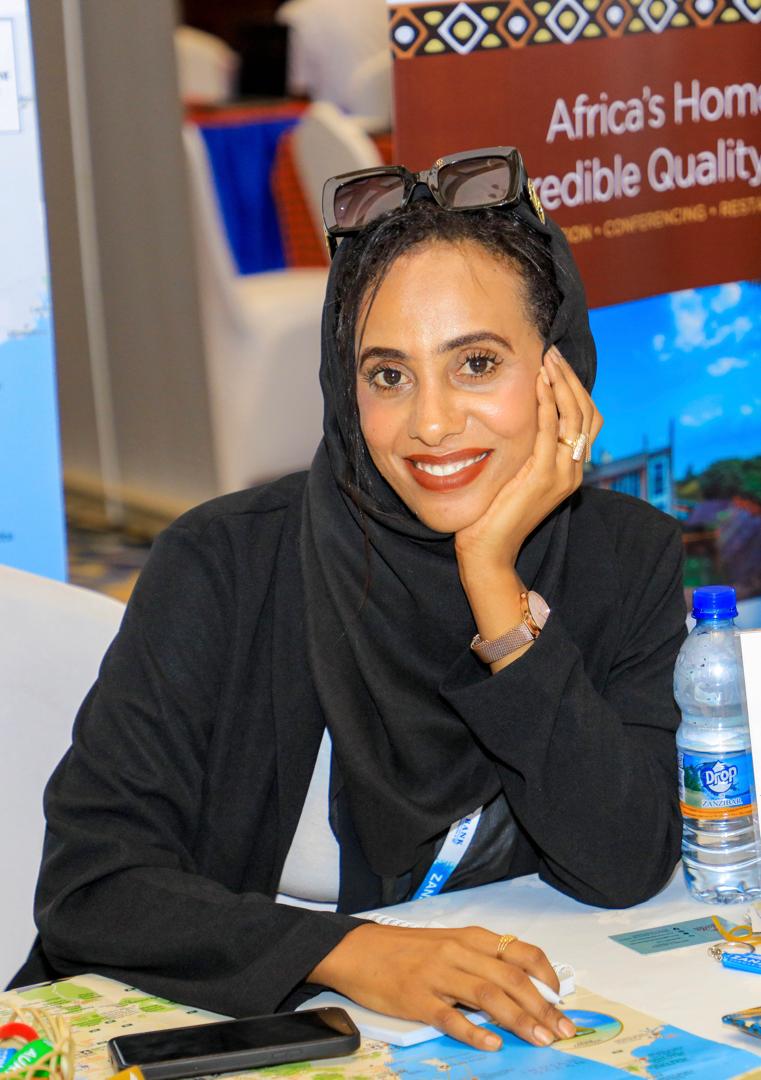 Smiling woman in a black hijab at a desk, with a water bottle and promotional materials around her.