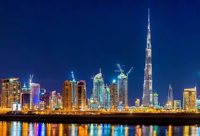 Night view of Dubai skyline with illuminated skyscrapers, including the Burj Khalifa, reflecting in the water.