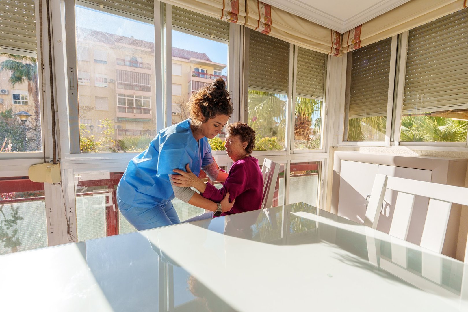 Young nurse is helping an elderly woman to stand up from a chair in a bright and airy room