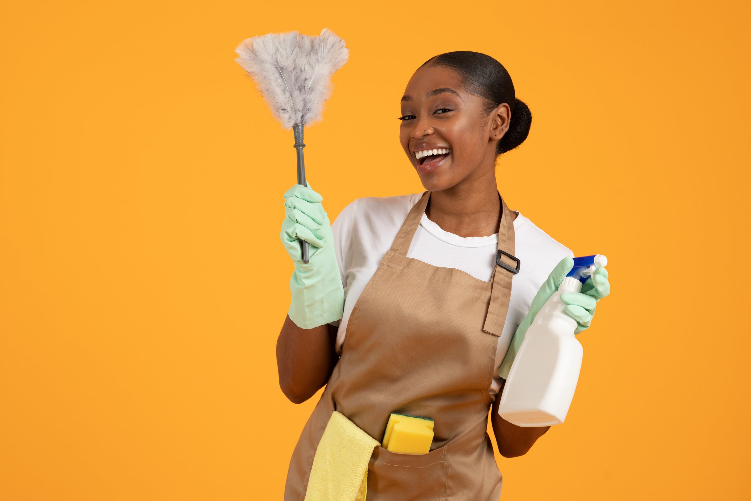 Portrait Of Joyful African American Lady Cleaner Holding Feather Duster And Detergent Spray, Enjoying Domestic Chores Over Yellow Backdrop In Studio, Advertising Professional Spring Cleaning Offer