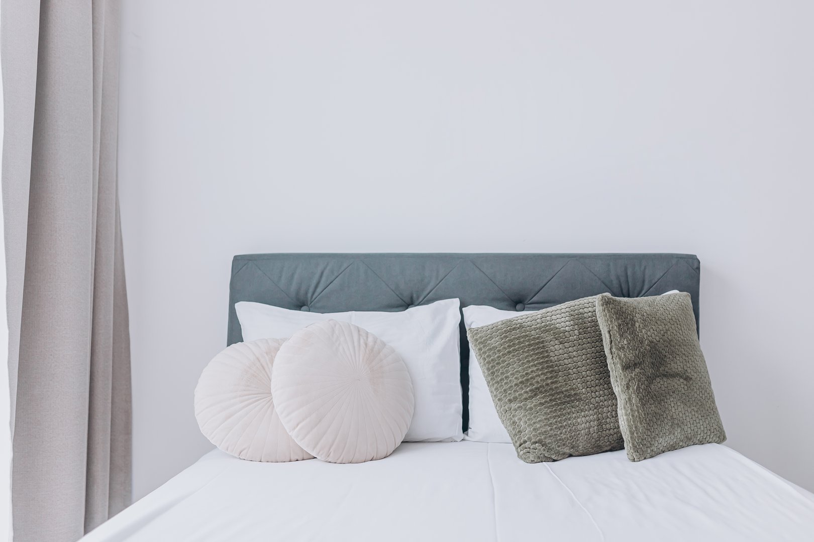 Simple bedroom interior with textured cushions, soft bedding, and fabric headboard. Beige curtain frames the space, emphasizing order and softness.