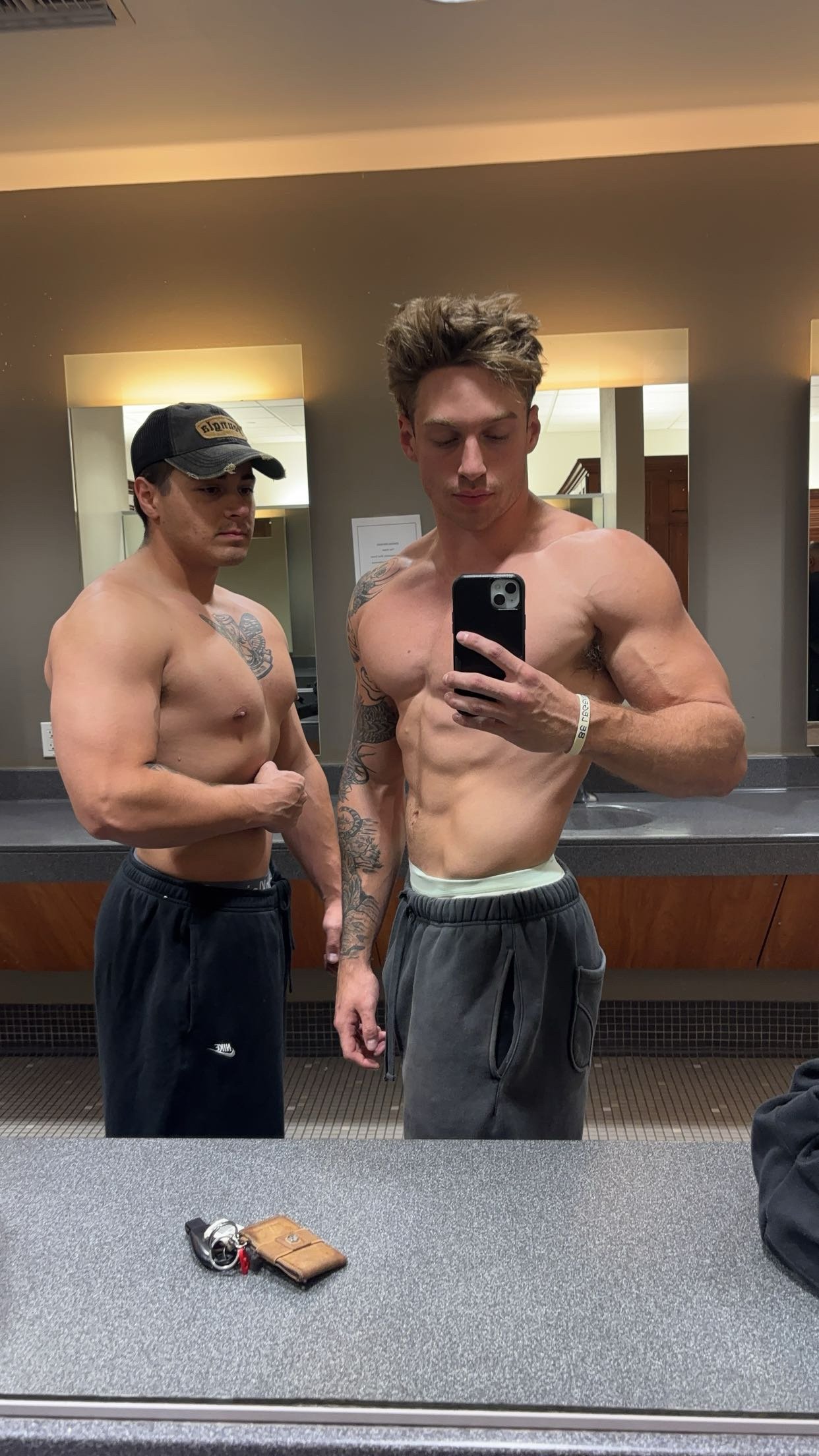 Two muscular men posing in a gym bathroom mirror, wearing casual gym attire. One holds a phone for a selfie.