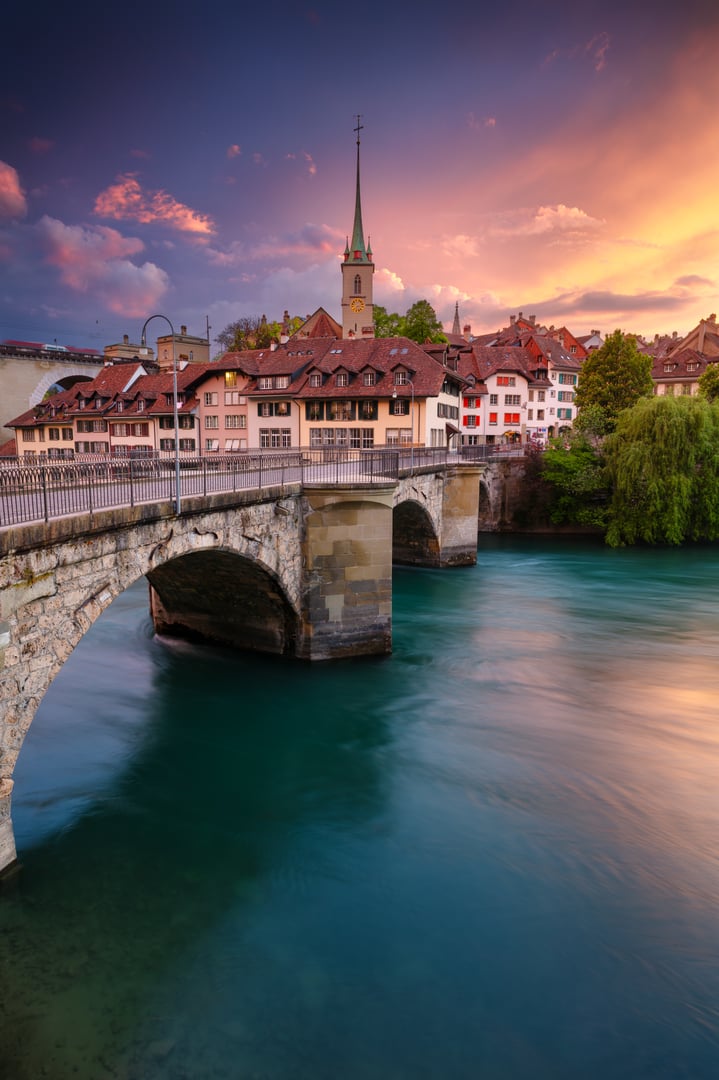 Arched stone bridge spans a river with teal water, leading to a charming cluster of traditional Swiss buildings. Gabled roofs and a slender church spire characterize the architecture, set against a vibrant, colorful sky at sunset. Lush green trees flank the scene, adding to the serene ambiance. The composition captures the essence of Bern, Switzerland, in a picturesque spring setting.