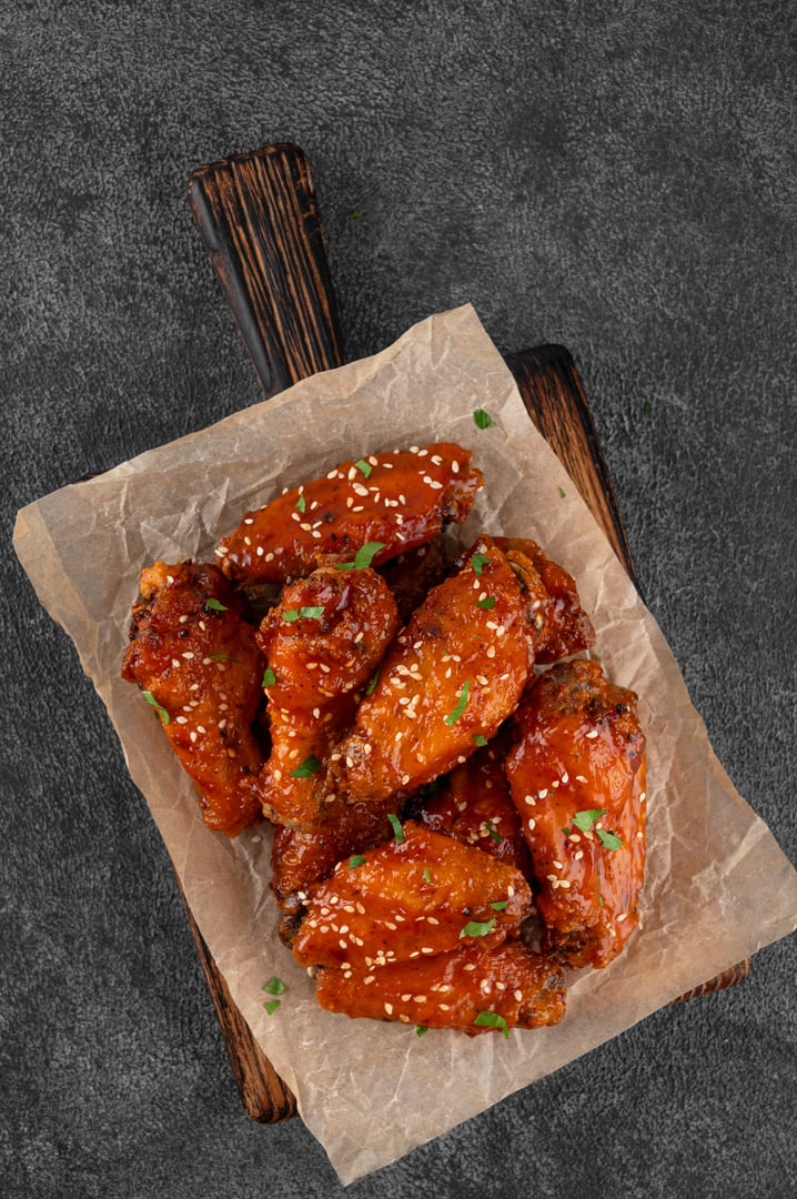 Baked chicken wings in a sweet chili sauce and sesame seeds on a wooden board on a dark concrete background. Top view, copy space