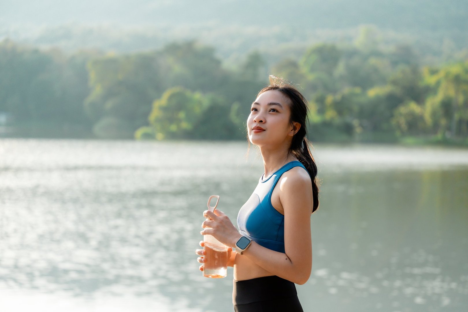 Young Asian sportswoman drinking water from bottle after training near lake in a sunny day