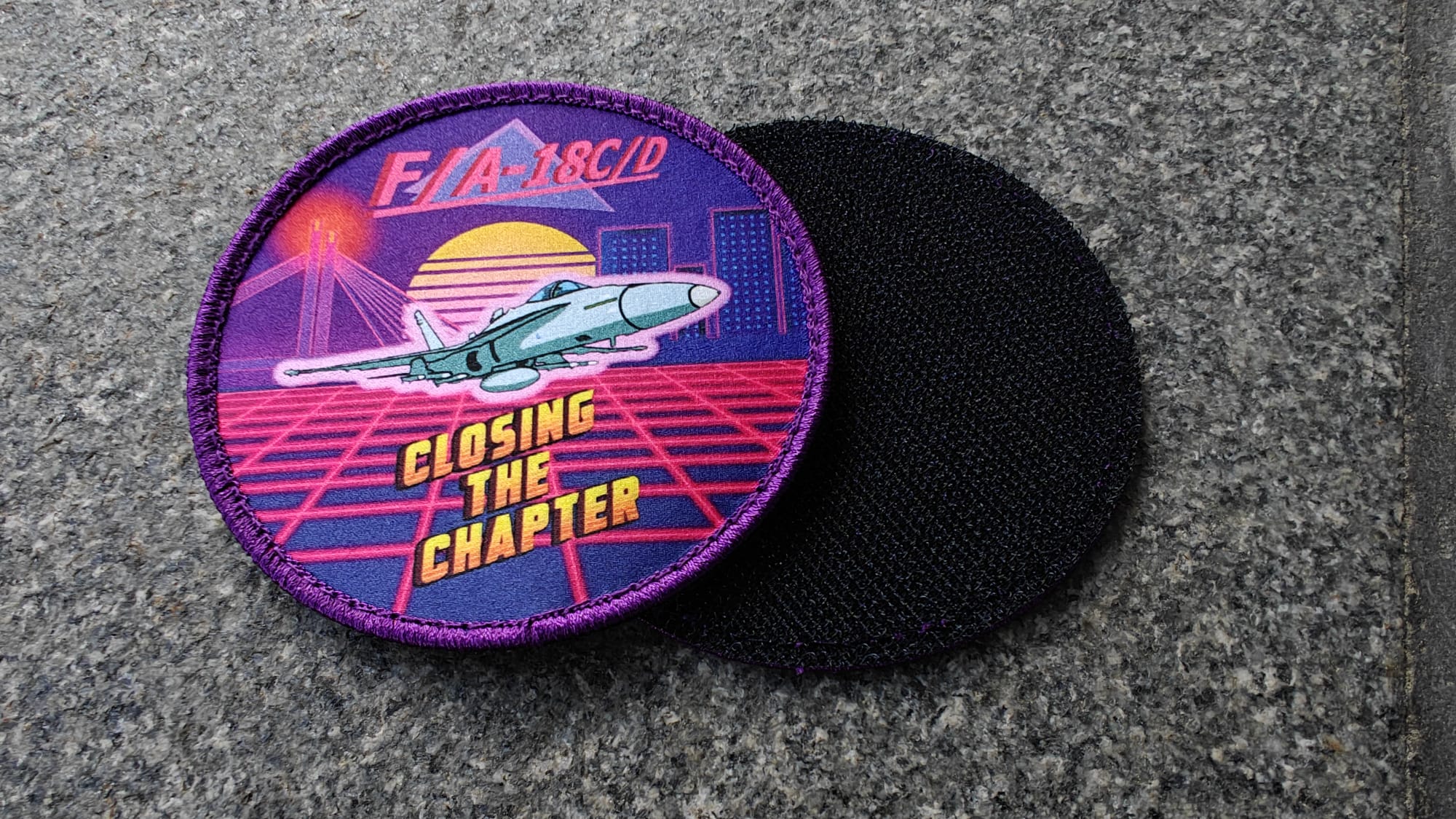 A colorful patch featuring an F/A-18C/D jet, a sunset, and the text Closing the Chapter, next to a plain black patch.