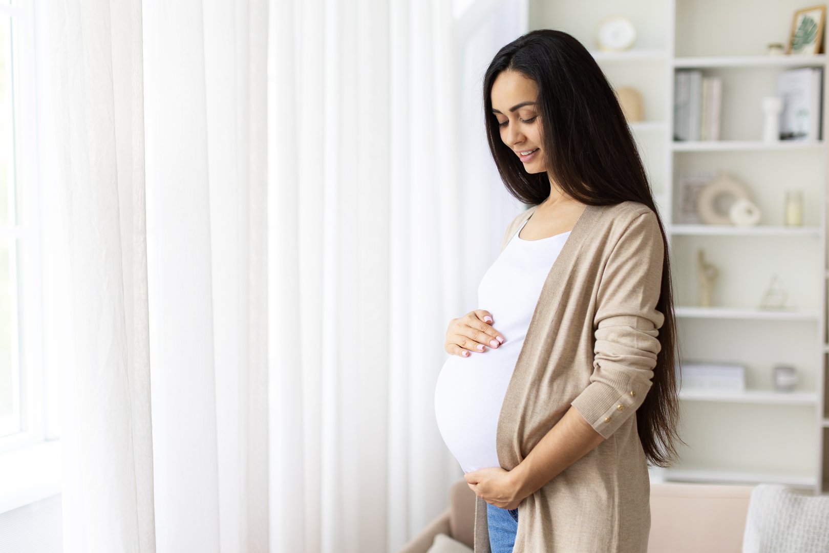 Pregnant young woman peacefully looking at belly while standing by the window. Calm atmosphere, cozy home and hopeful moment captured in bright natural lighting, copy space