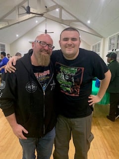 Two men smiling and posing together in a brightly lit room with wooden floors and high ceilings.