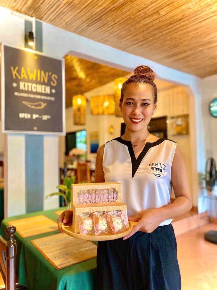 A woman holding a tray with pastries, smiling inside a cafe with a sign reading Kawins Kitchen.