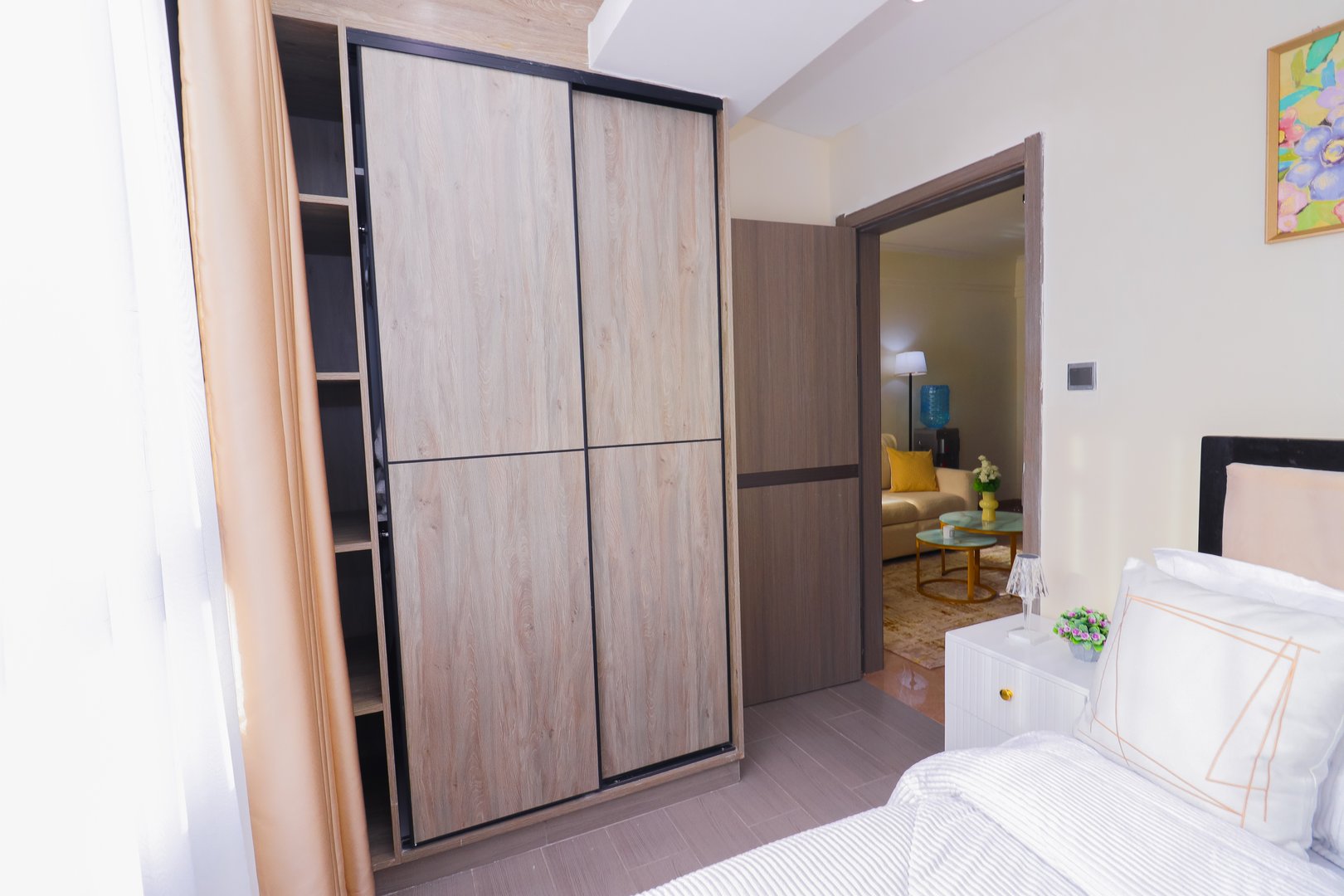 Modern bedroom with wardrobe, open sliding door revealing a living room, neutral tones, and minimal decor.